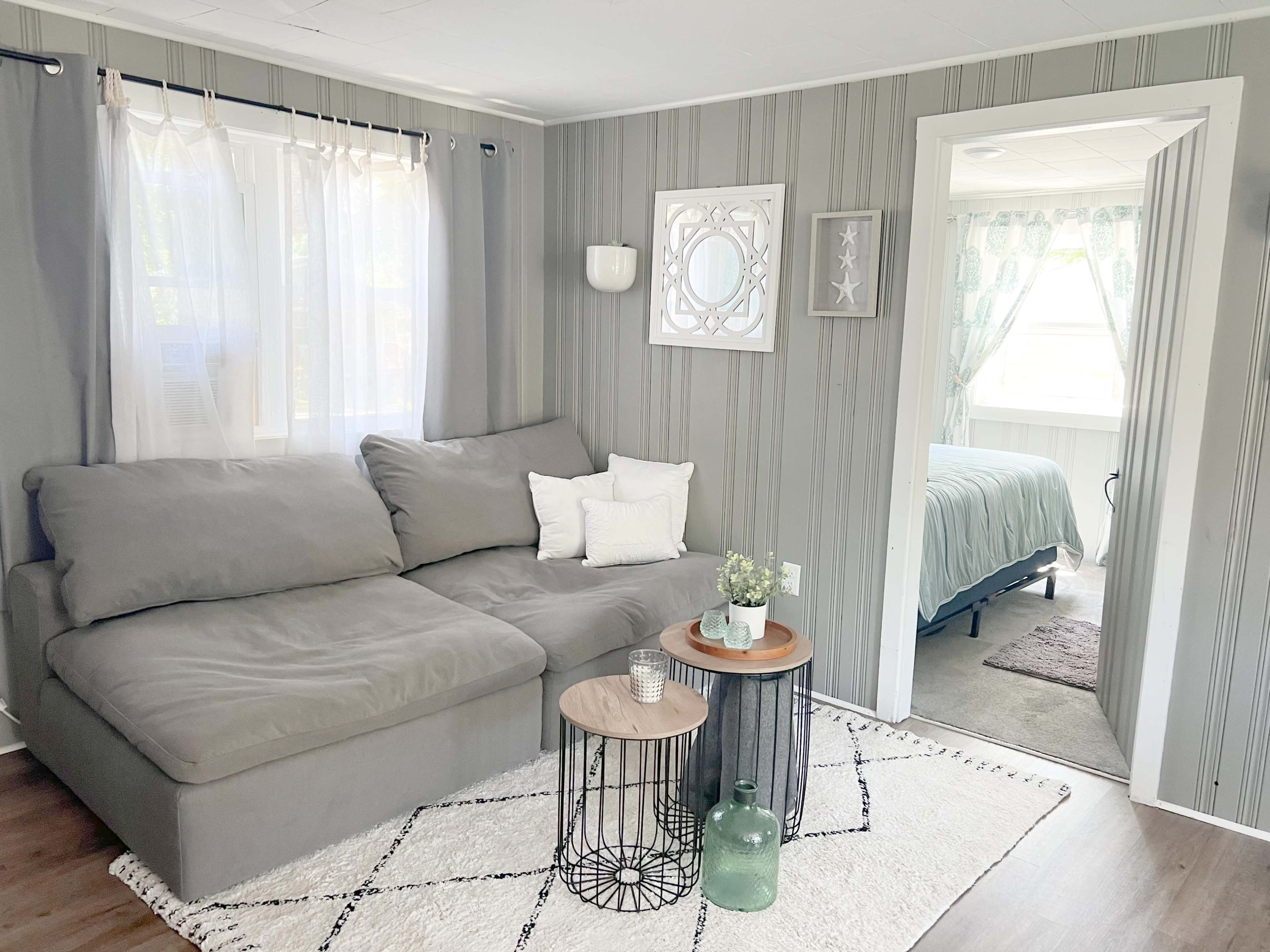 The image shows a cozy living room with a gray sectional sofa, decorative pillows, two side tables, and a doorway leading to a bedroom area.