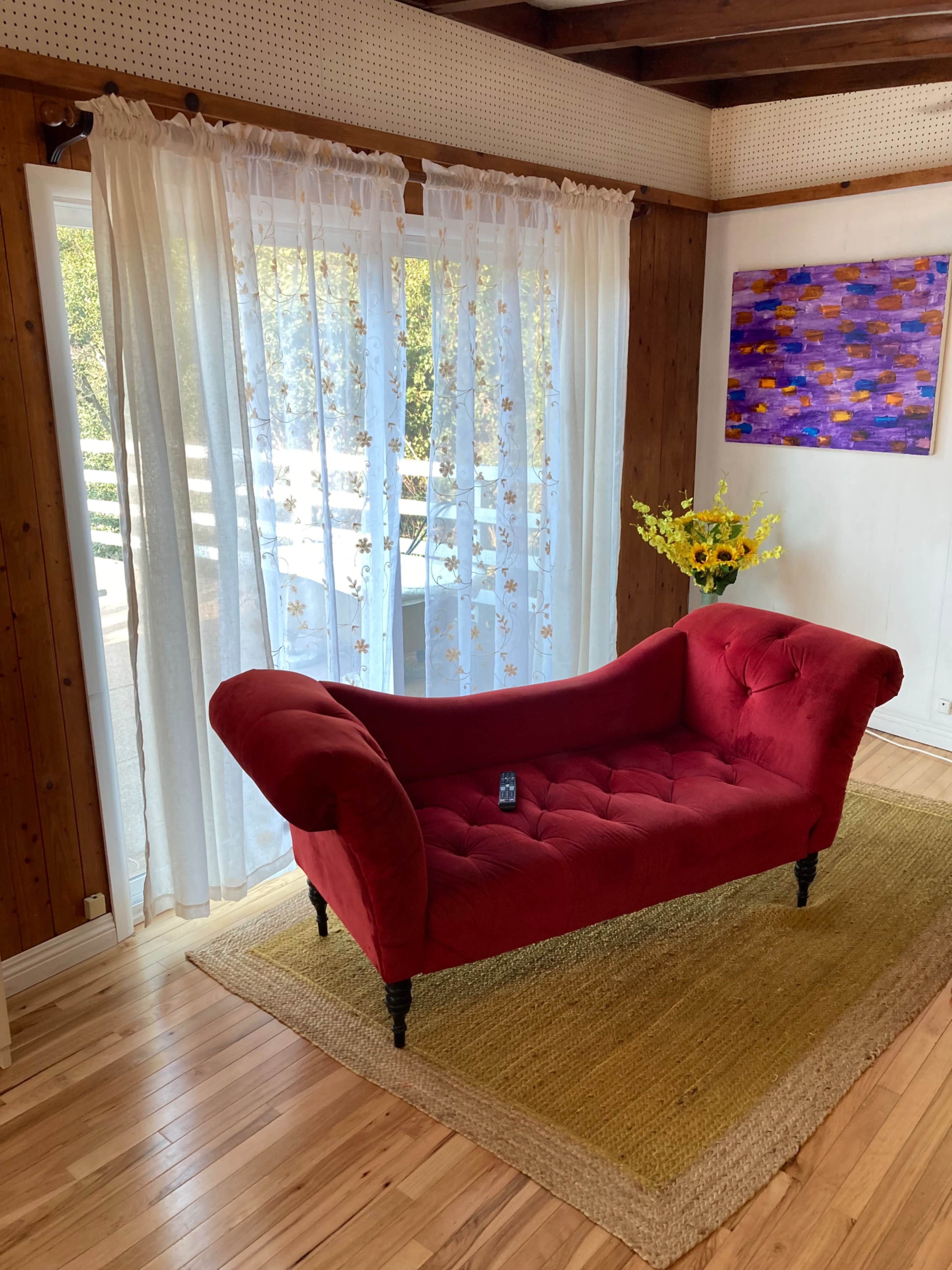 A red tufted couch is positioned on a light brown area rug in a room with wooden walls and large curtains letting in natural light.