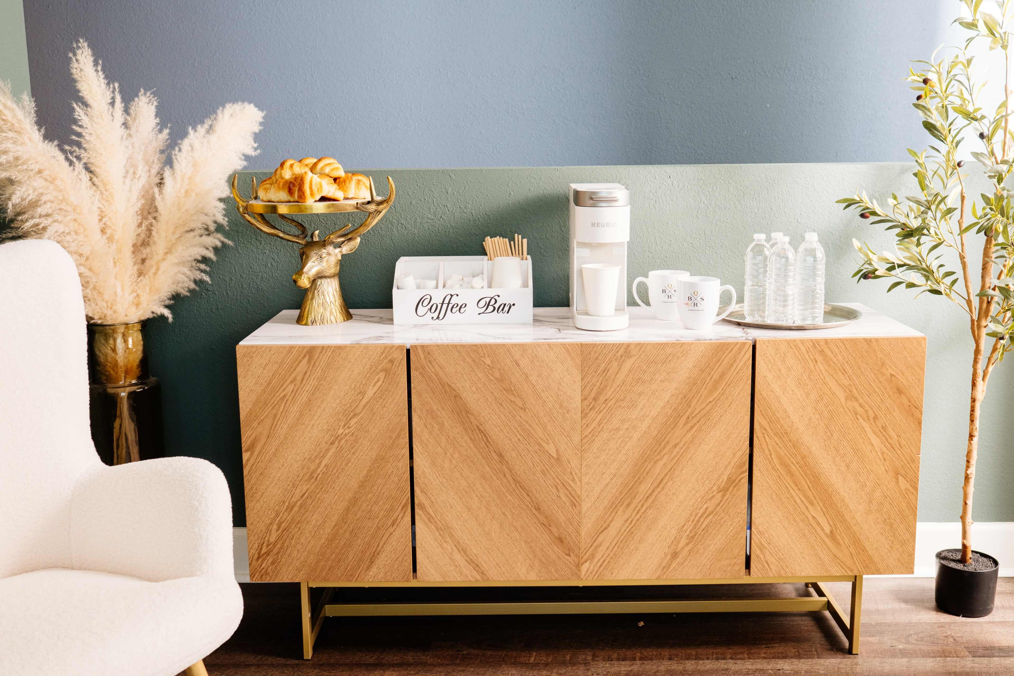 A wooden sideboard features a coffee bar setup with pastries, water bottles, and a coffee maker against a painted wall.