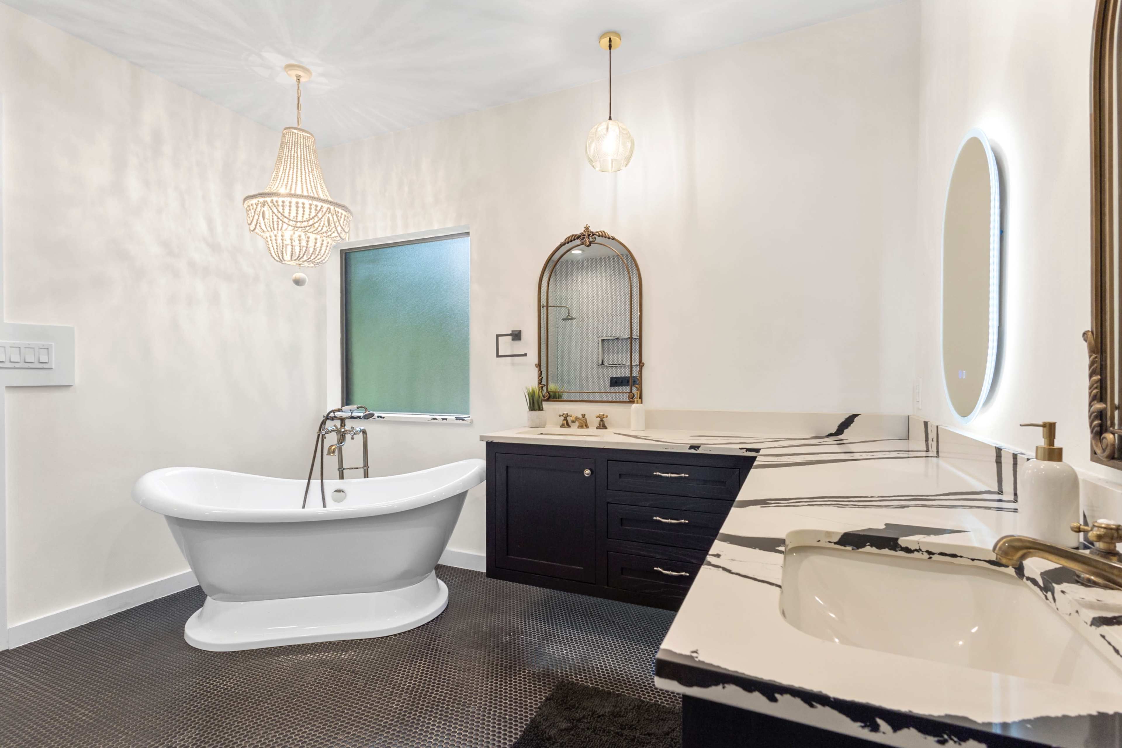 The image shows a modern bathroom featuring a freestanding white bathtub, a dual-sink vanity with a black countertop, and elegant lighting fixtures.