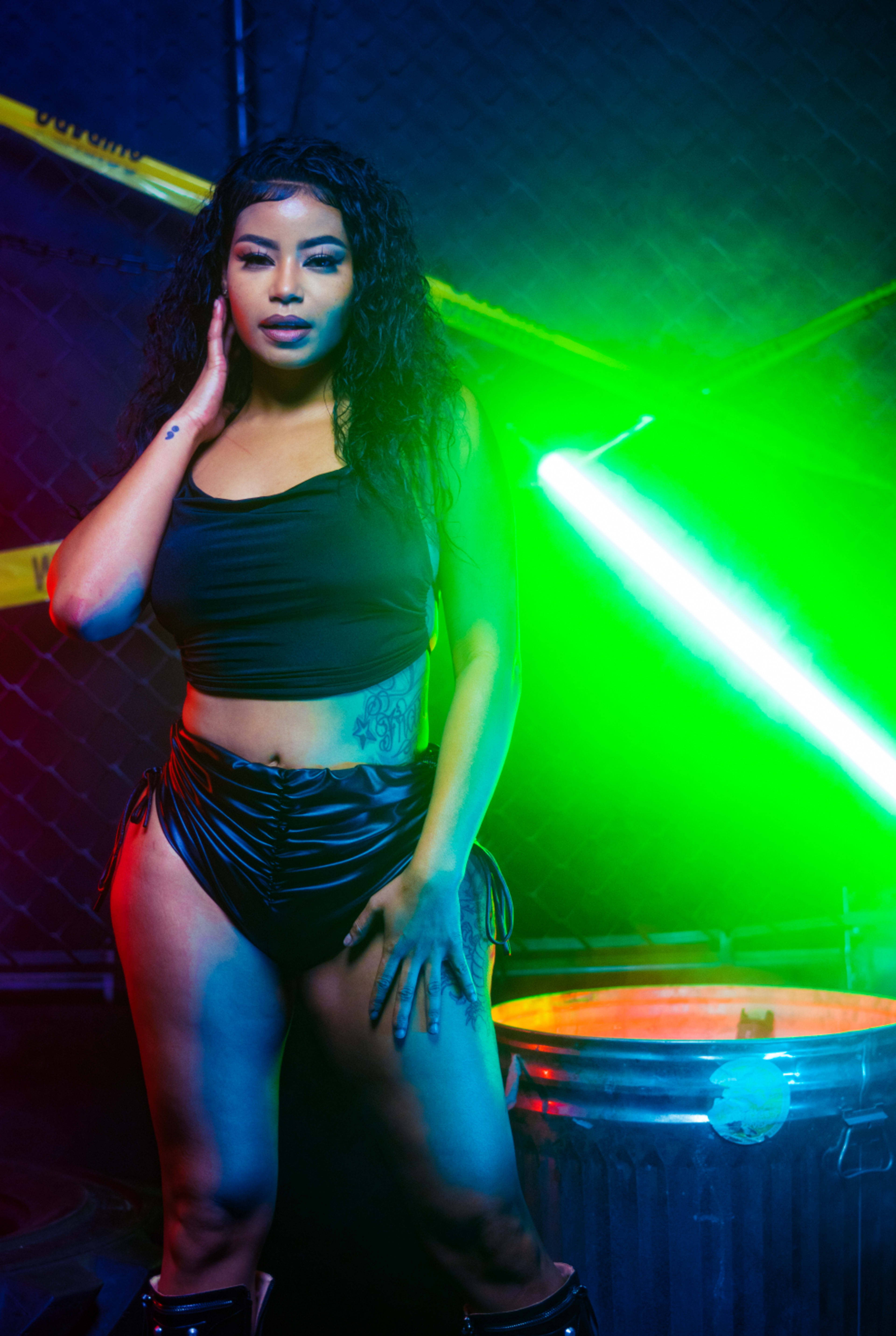 A woman poses confidently in a dimly lit space, featuring colorful lighting and a metal trash can in the background.