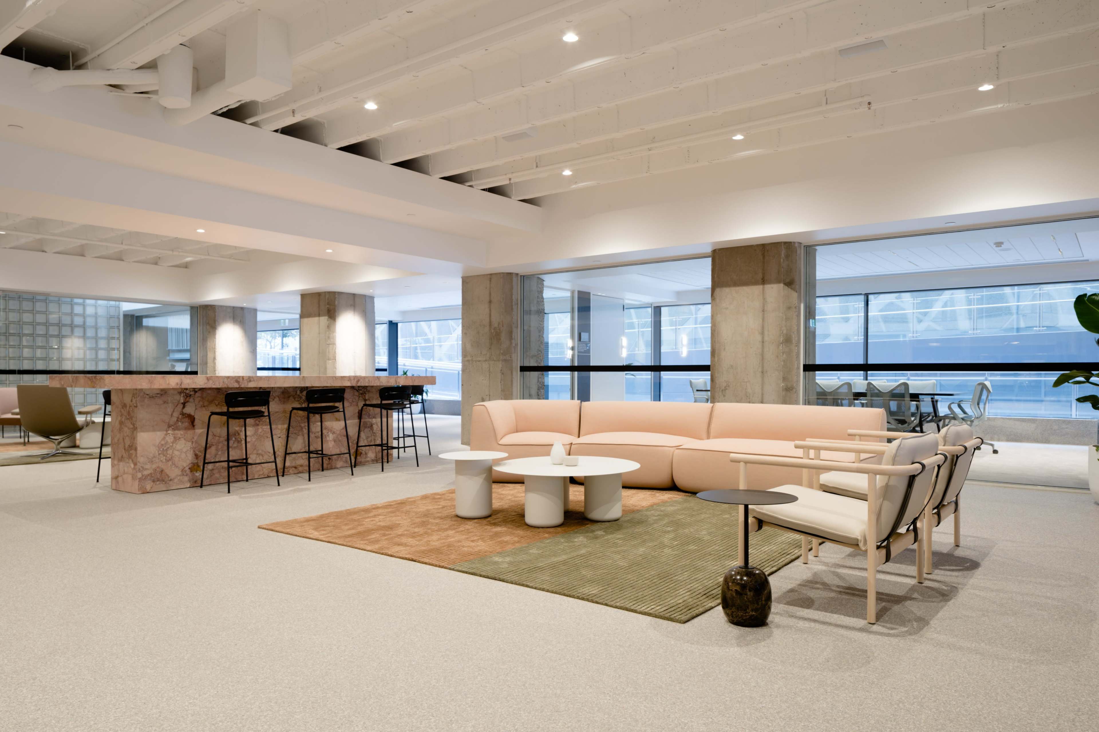 A modern office lounge area featuring a pink sectional sofa, a marble bar with high stools, and various tables arranged on a light-colored carpet.