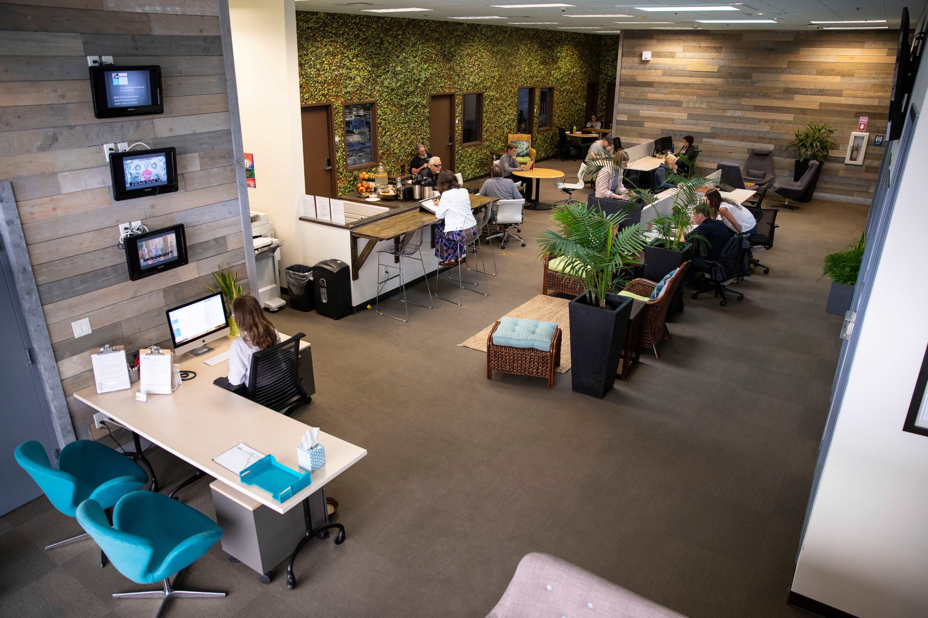 The image shows a modern office workspace with multiple workstations, a communal seating area, and plants along the walls.