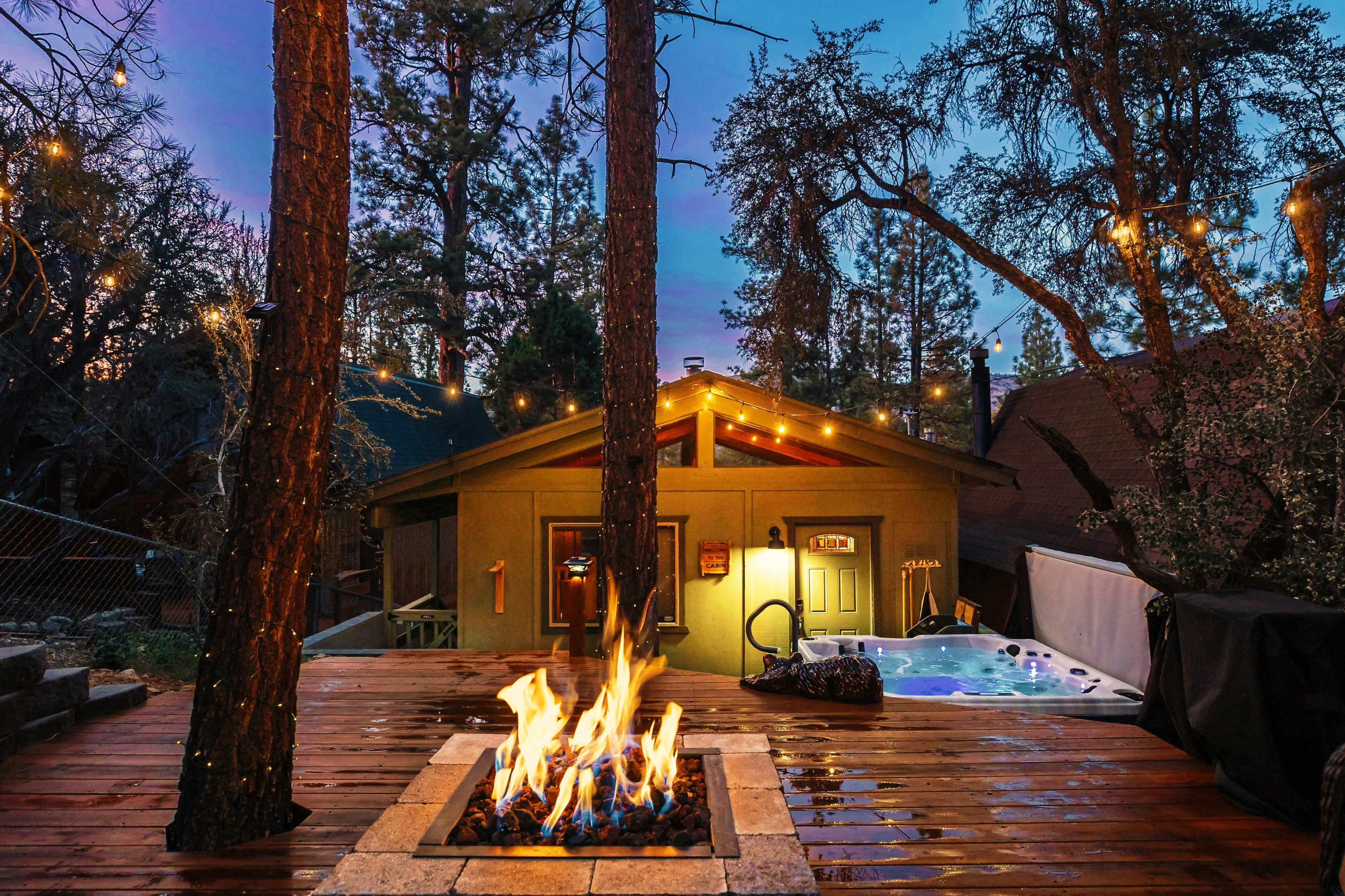 A fire pit surrounded by wooden decking and trees is located near a hot tub outside a cabin, illuminated by string lights at dusk.