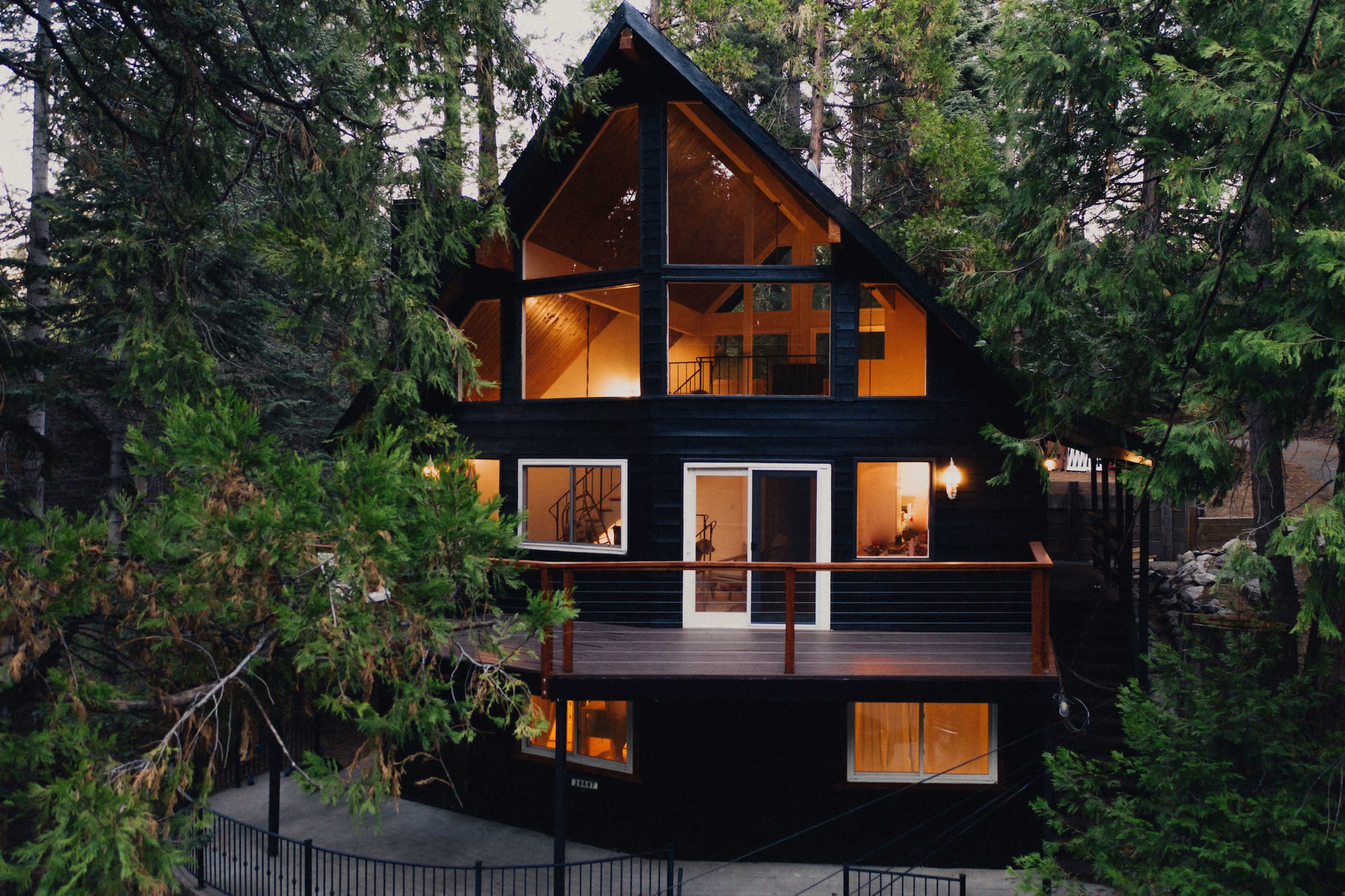 A modern black cabin with large windows sits amidst tall green trees.