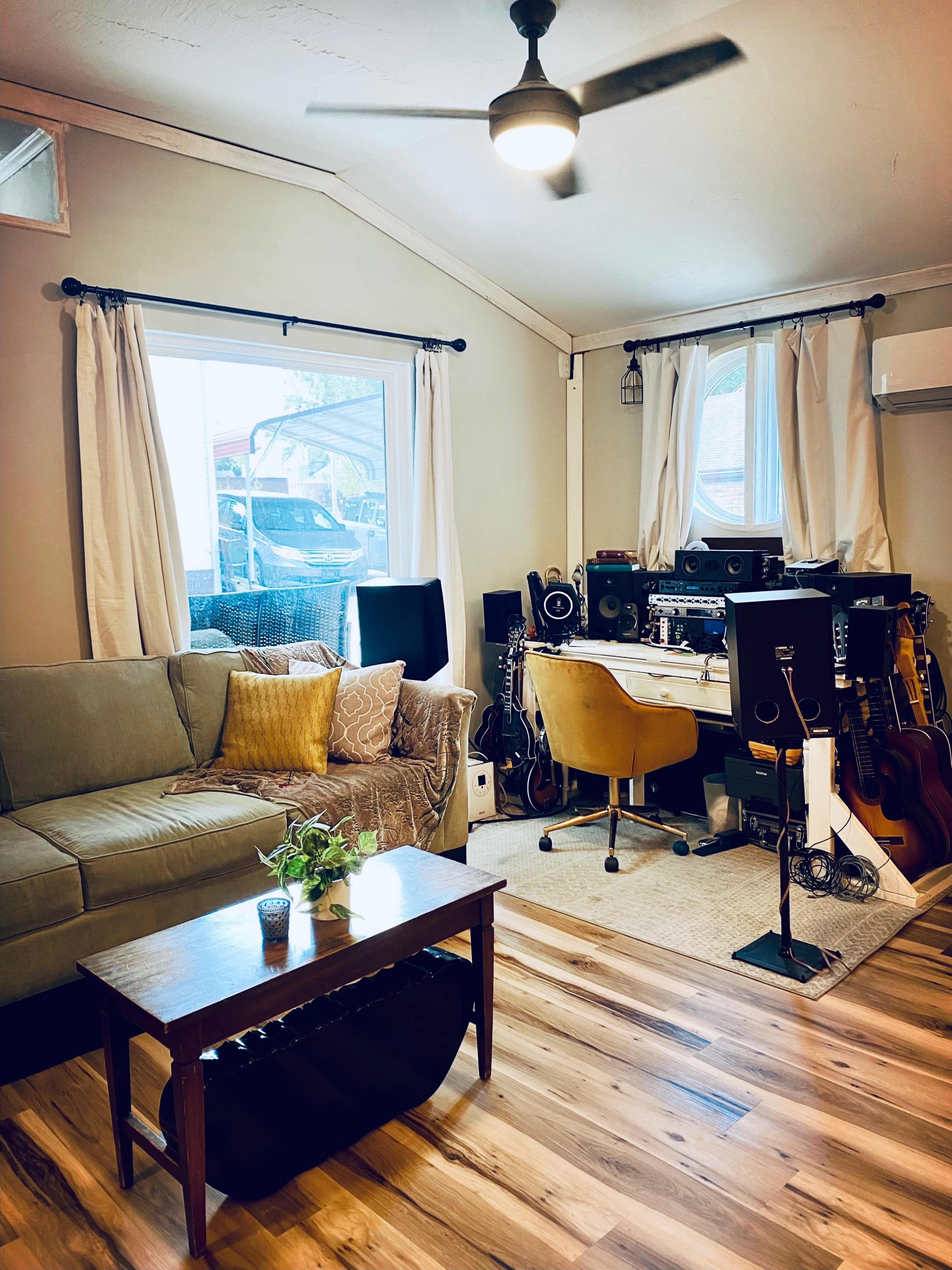 The image shows a cozy living room with a green couch, a wooden coffee table, and a workspace equipped with audio equipment and musical instruments.