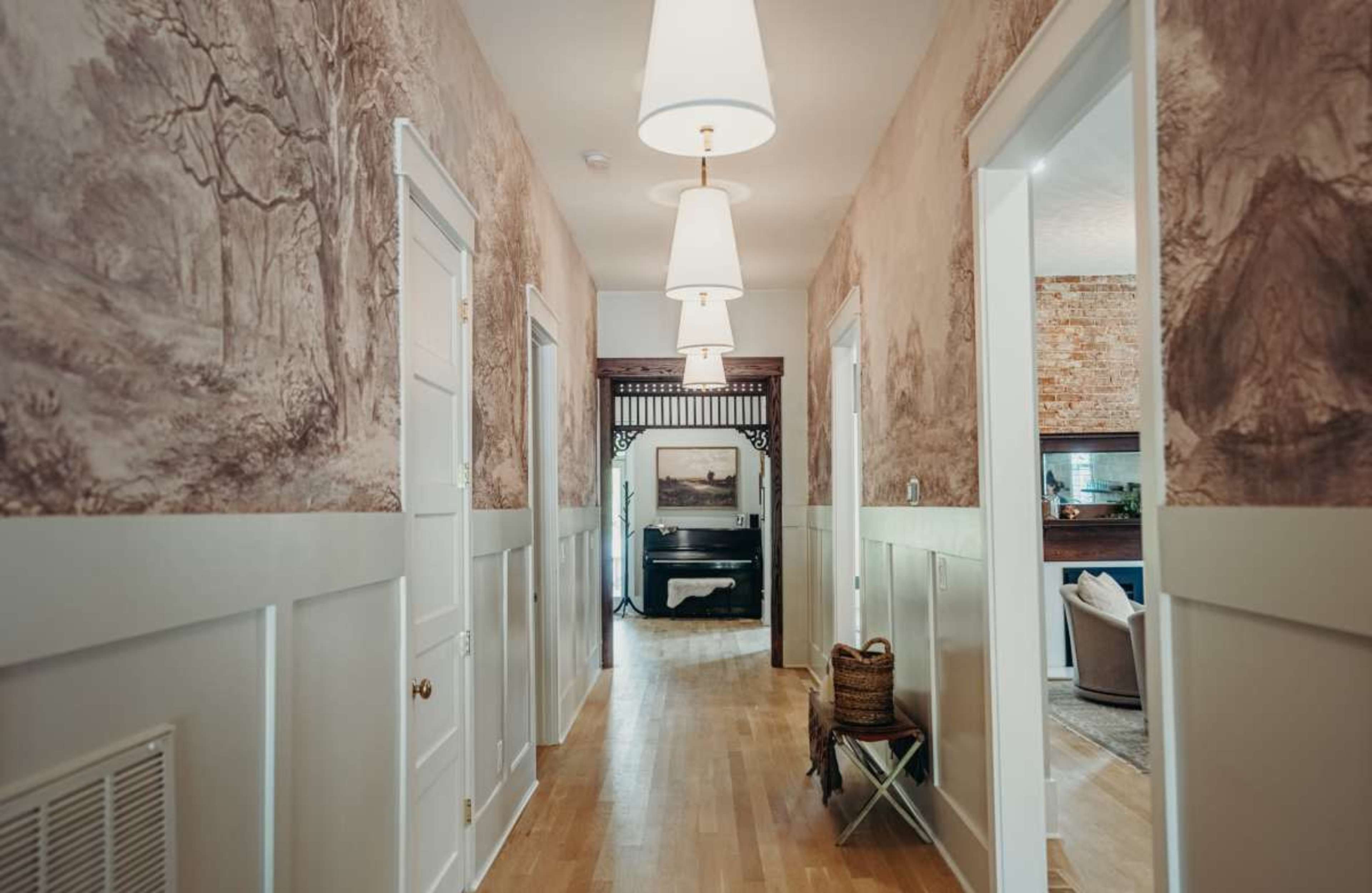 The image shows a hallway with light-colored walls featuring mural wallpaper, lit by pendant lights hanging from the ceiling, and doors on either side leading to rooms.