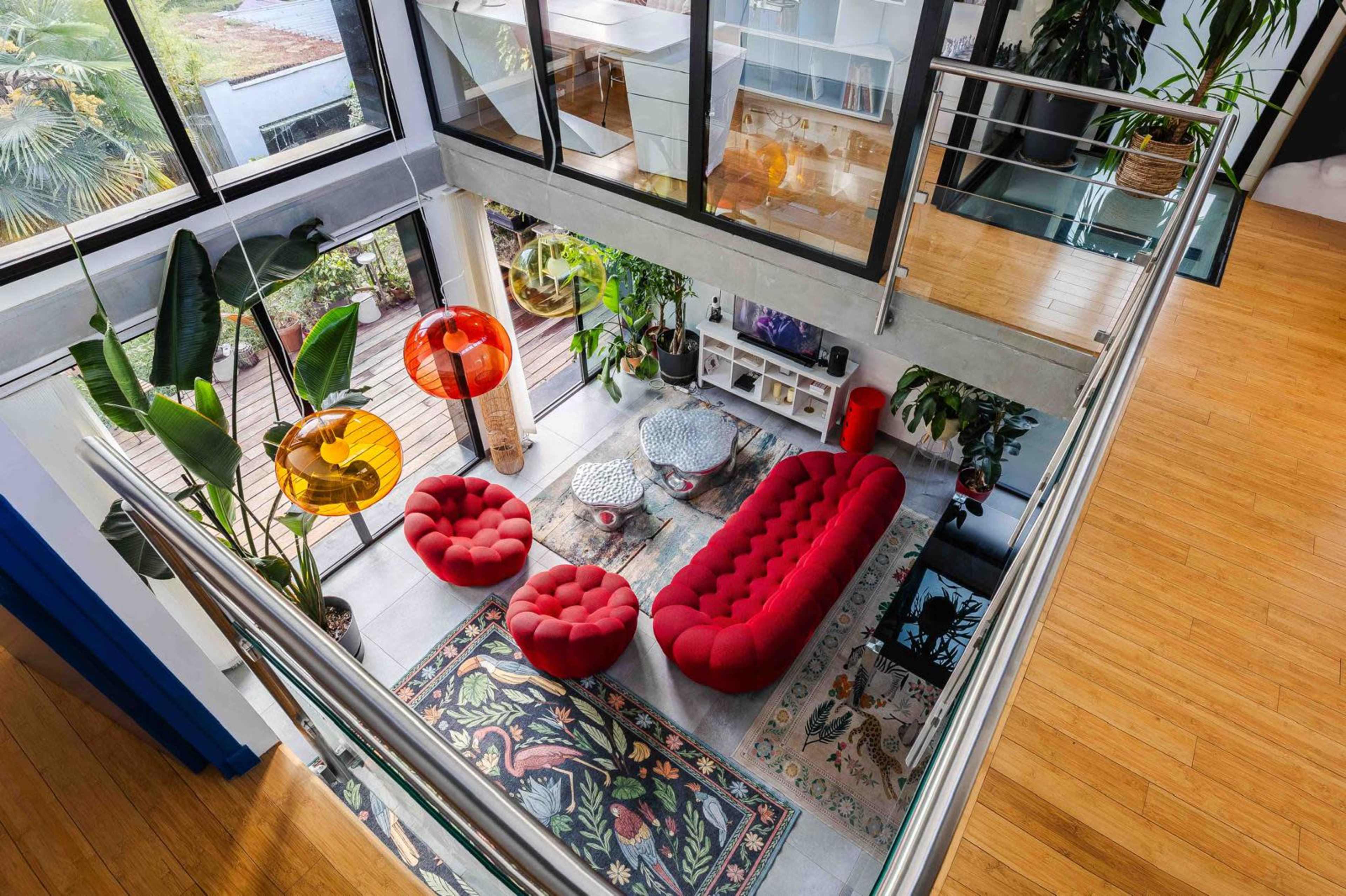 The image shows a modern living room with a glass railing that overlooks a space featuring red furniture, decorative rugs, and various houseplants.