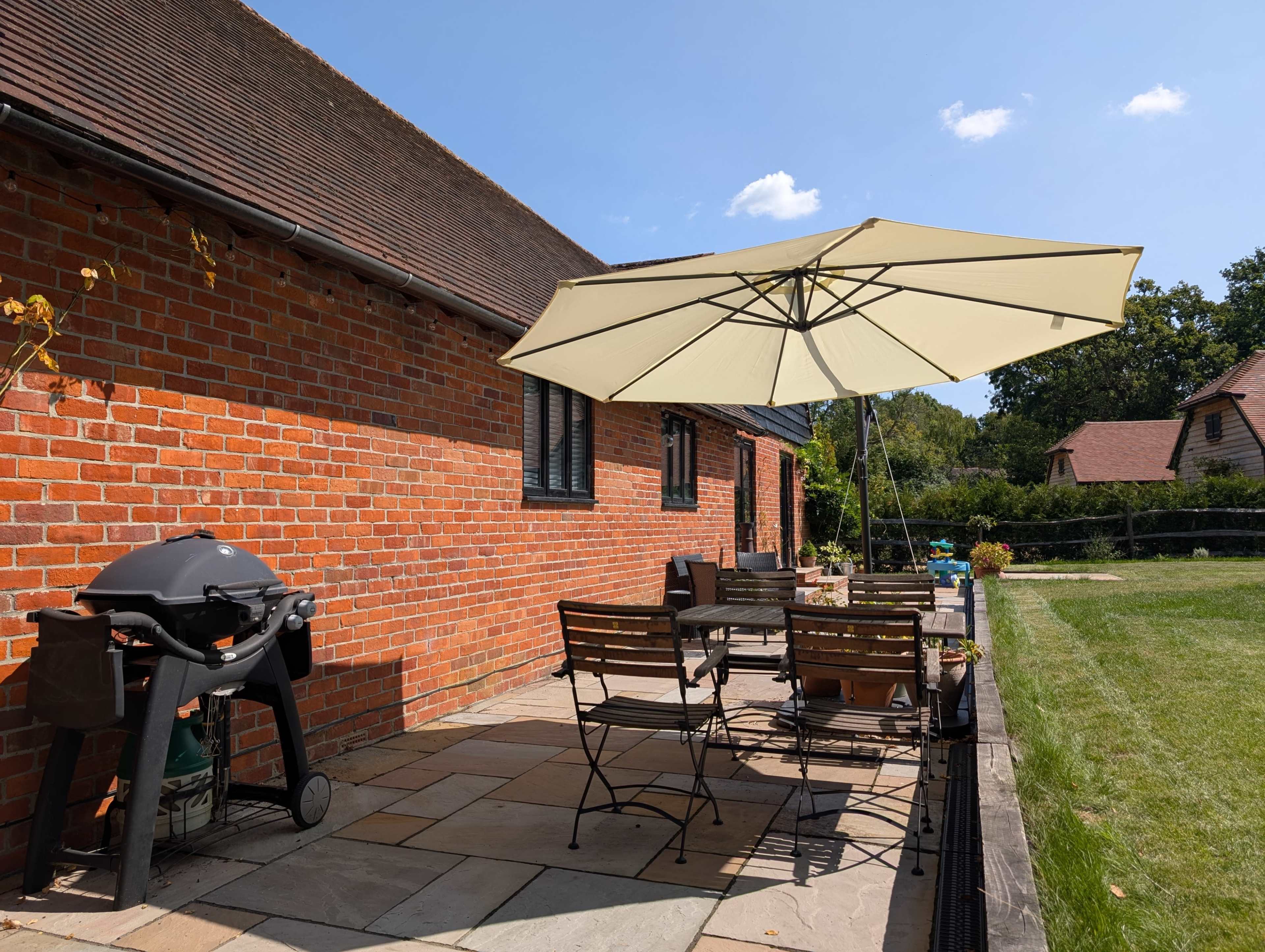 A patio features a dining set with chairs and a table, an umbrella for shade, and a grill, situated against a brick wall of a house.