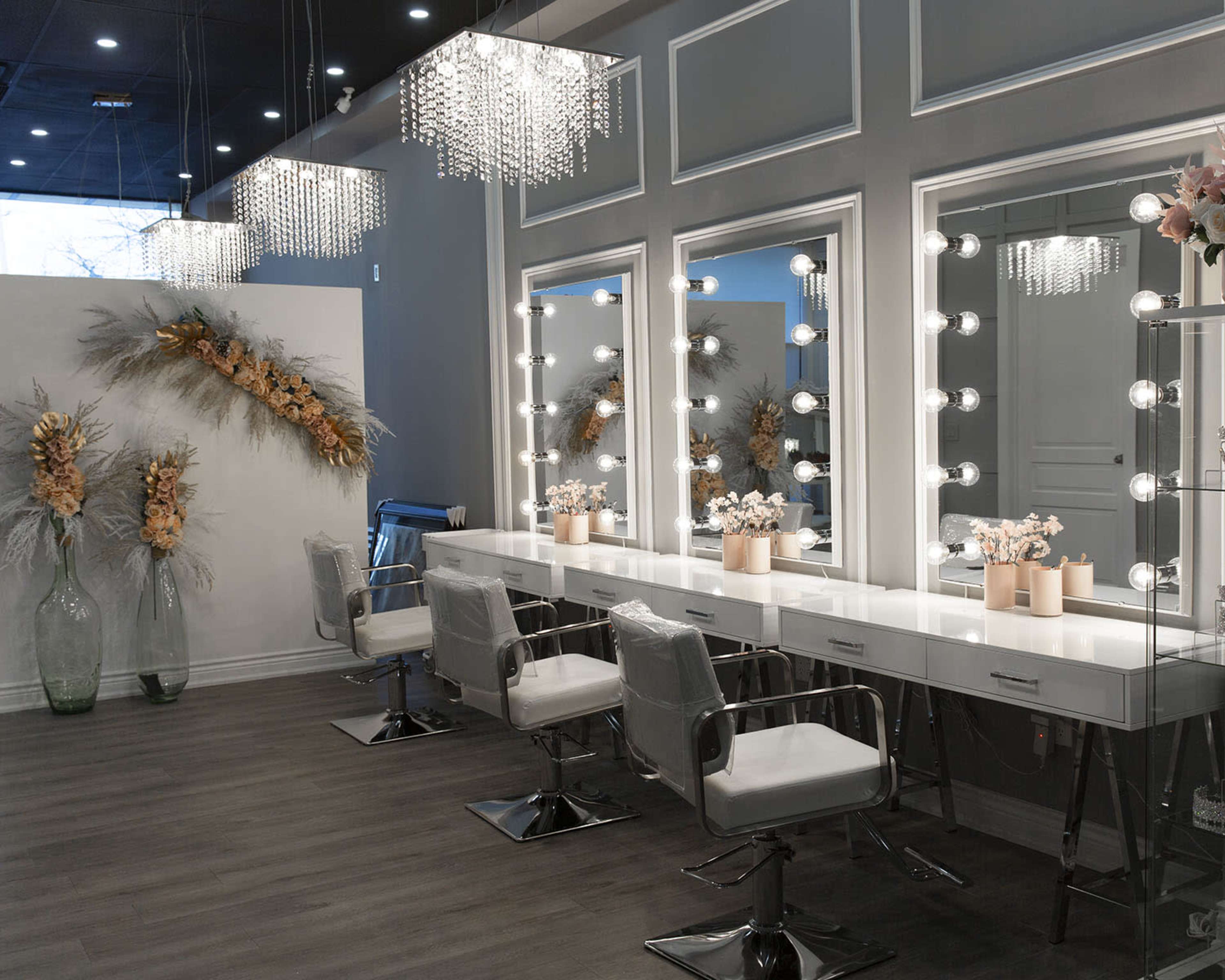 A modern makeup studio featuring three illuminated mirrors above a row of white vanities with stylish chairs and decorative floral arrangements.