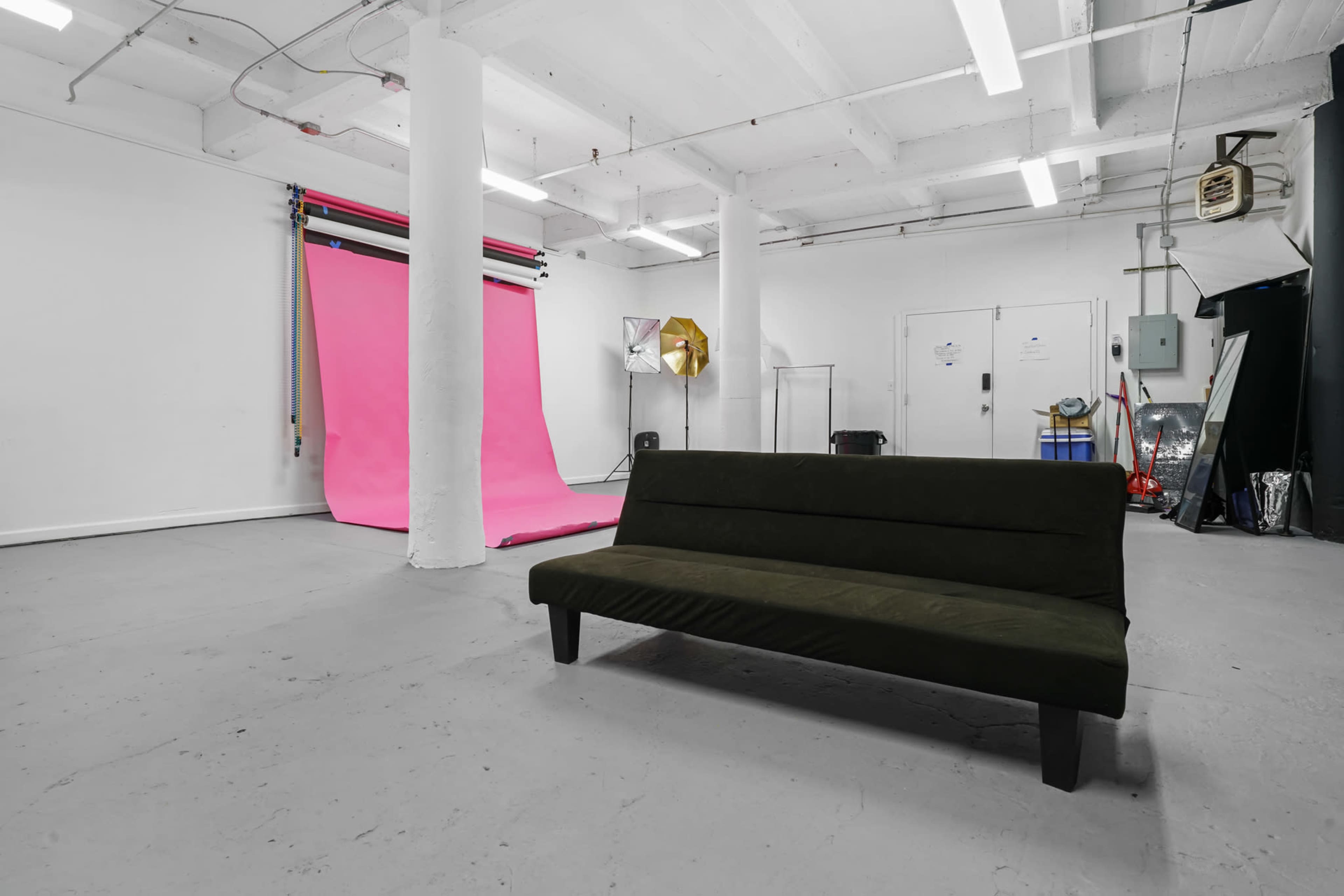 A minimalistic studio space with a green sofa in the foreground and a vibrant pink backdrop behind it, accompanied by various lighting equipment.
