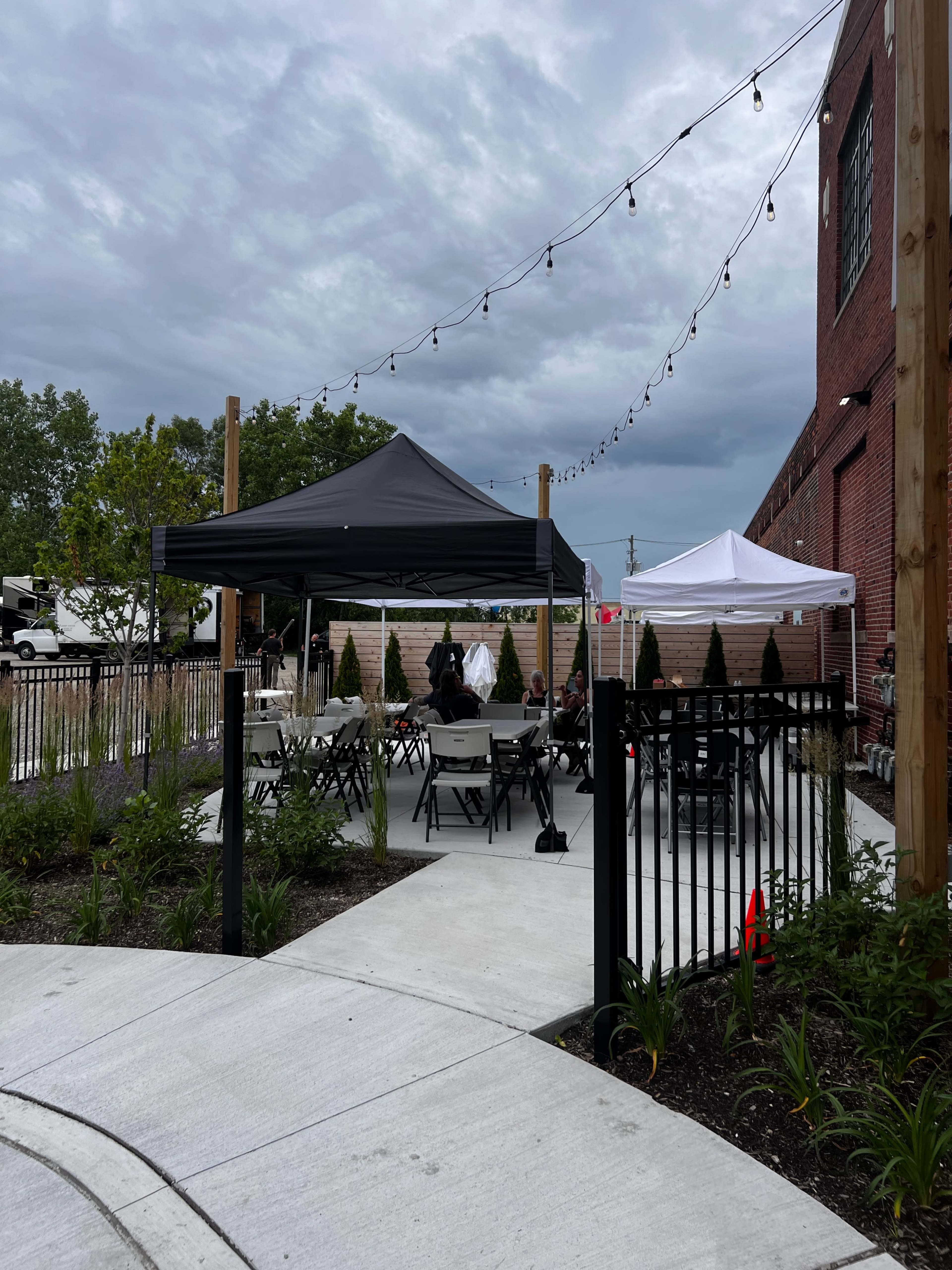 A patio area is set up with black and white tents, surrounded by tables and chairs, under a cloudy sky with string lights overhead.