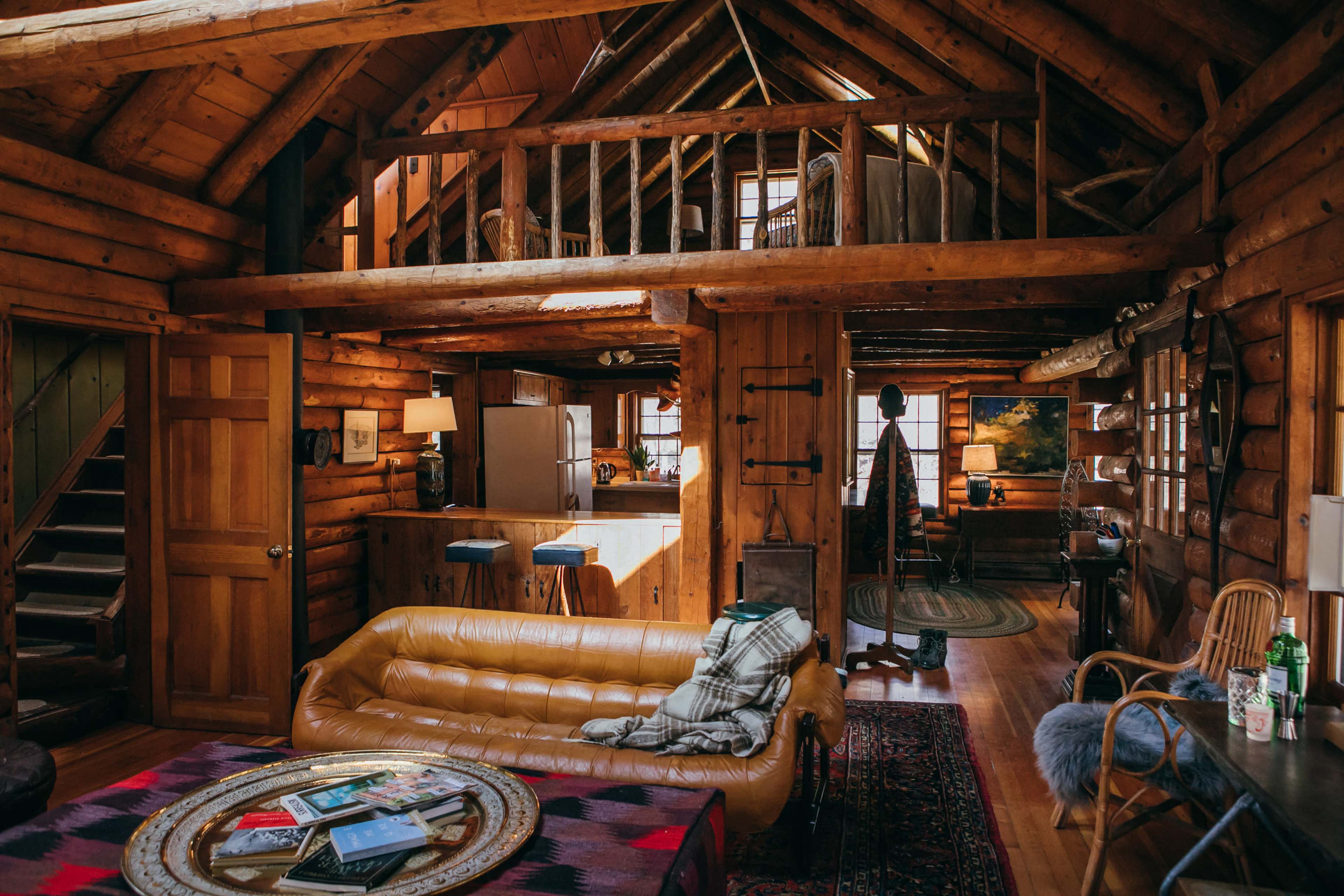 The interior of a rustic log cabin features exposed wooden beams, a lofted sleeping area, and a cozy living space with a brown leather sofa and a staircase leading to the upper level.