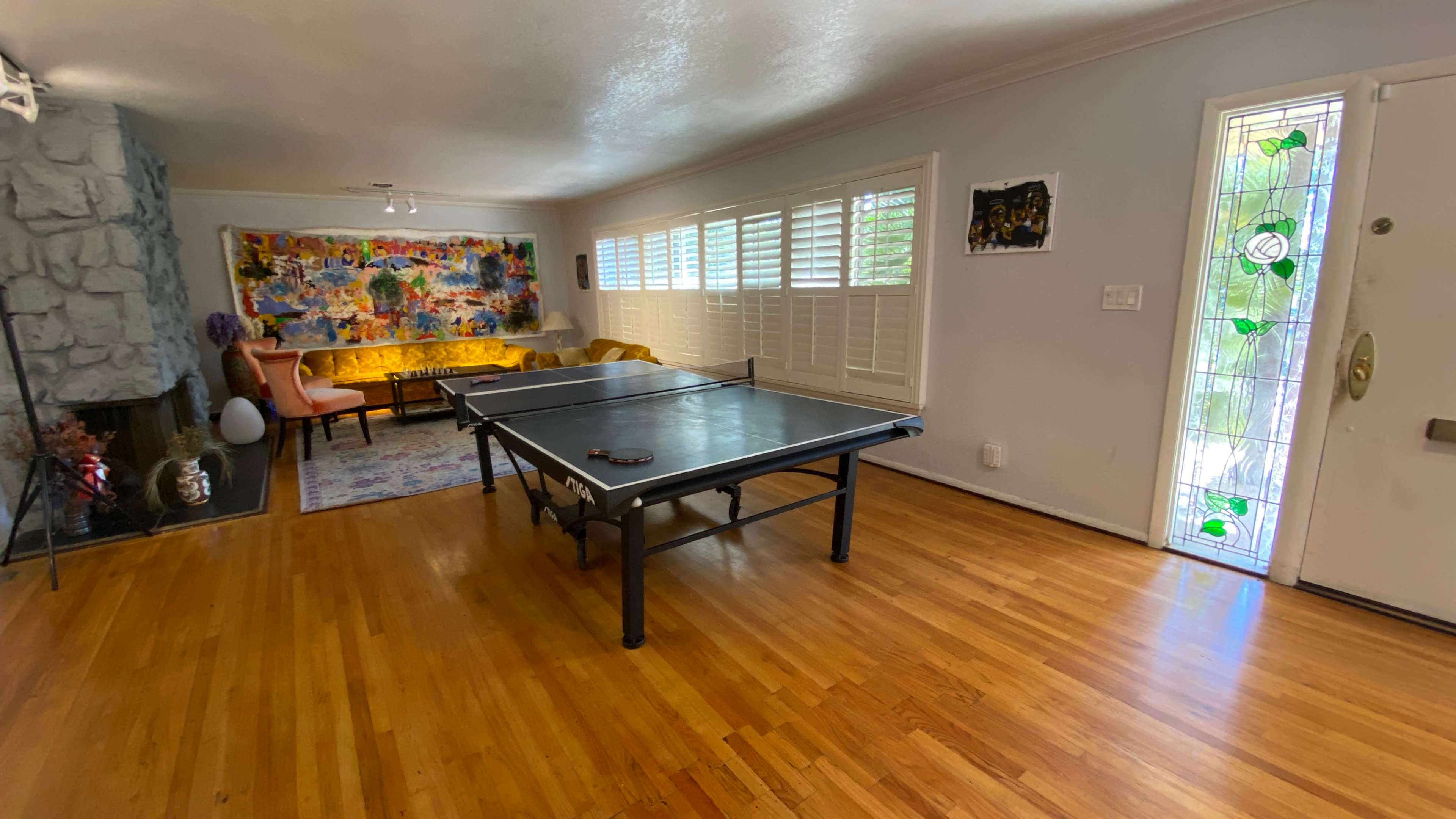 The image shows a living room with a ping pong table in the center, hardwood flooring, a colorful wall mural, a sofa, and a front door with stained glass.