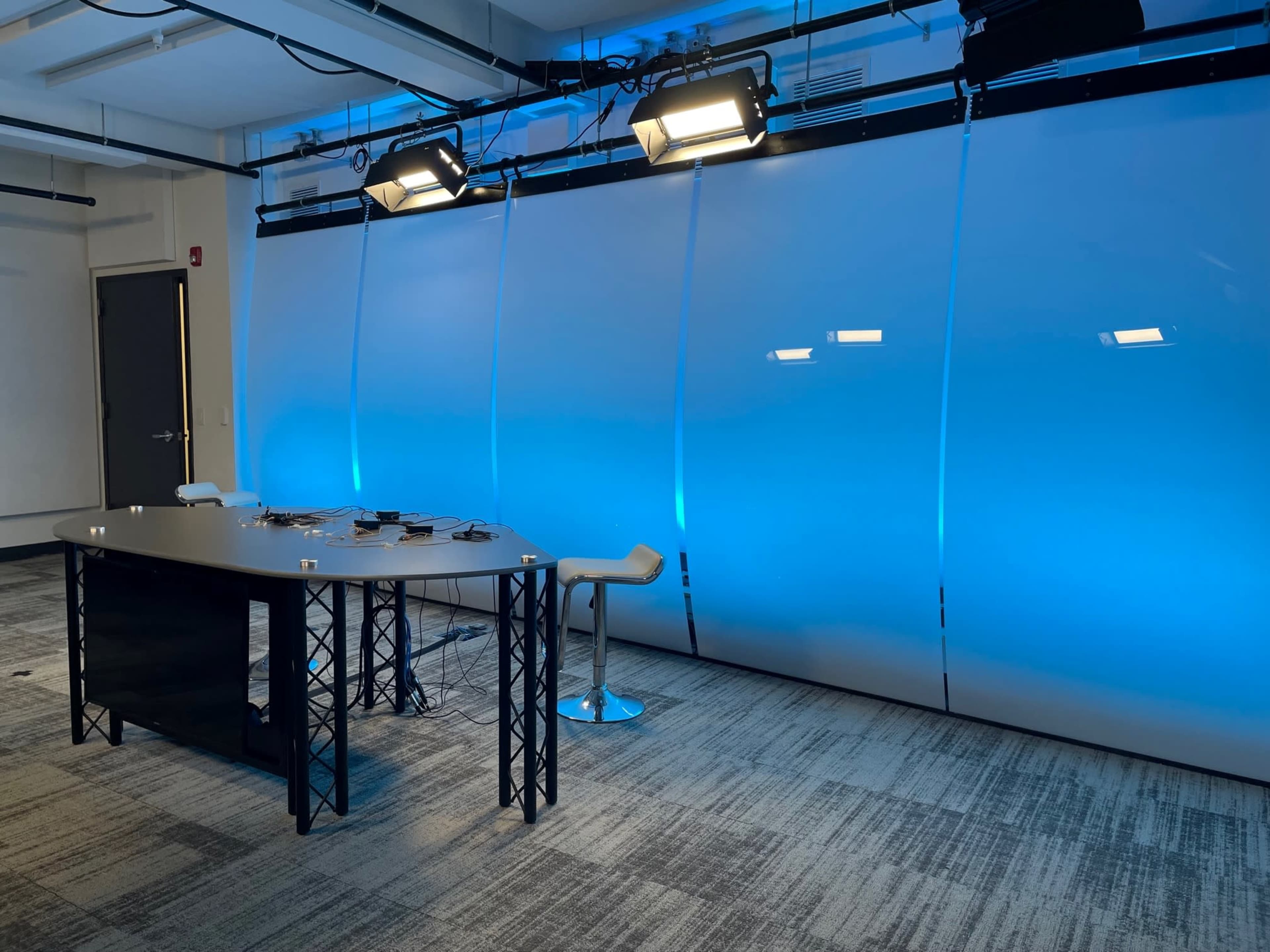 A modern workspace features a sleek table, stools, and blue-illuminated backdrop with overhead lighting.