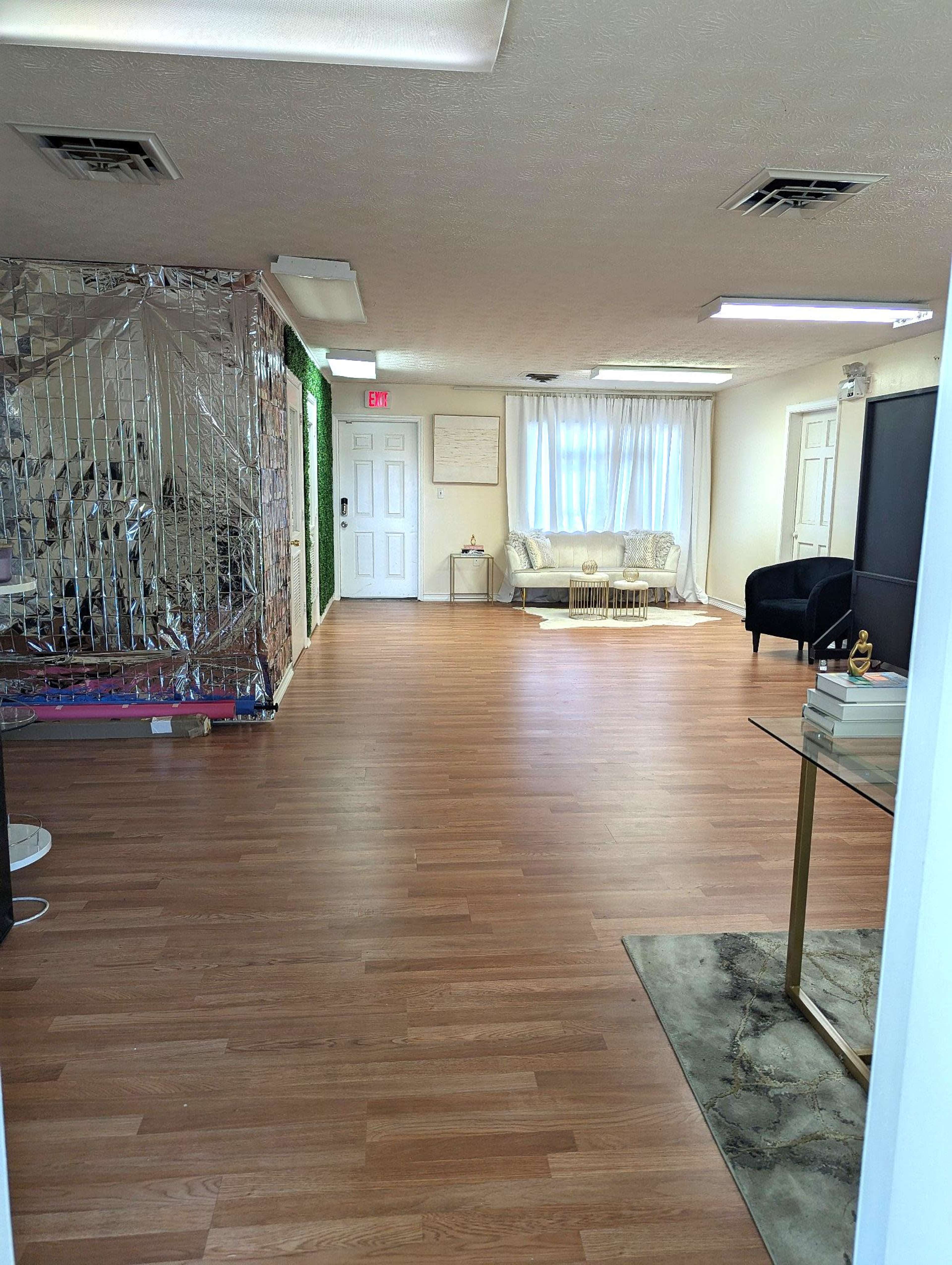 The image shows a spacious room with wooden flooring, a large window covered by sheer curtains, a couch, and a partially mirrored wall.