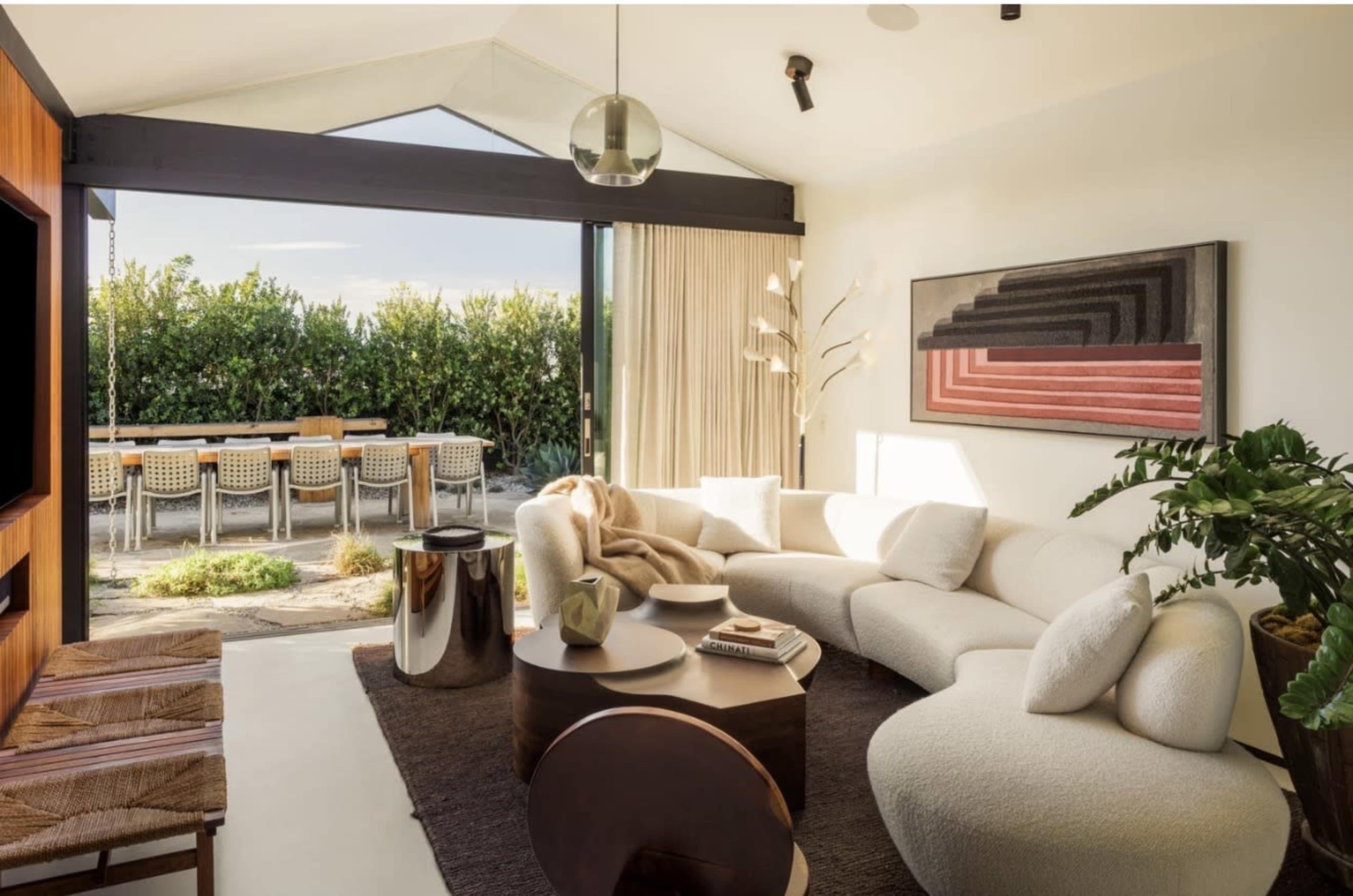 The image shows a modern living room with a curved white sofa, a round coffee table, and large glass doors leading to an outdoor dining area surrounded by greenery.