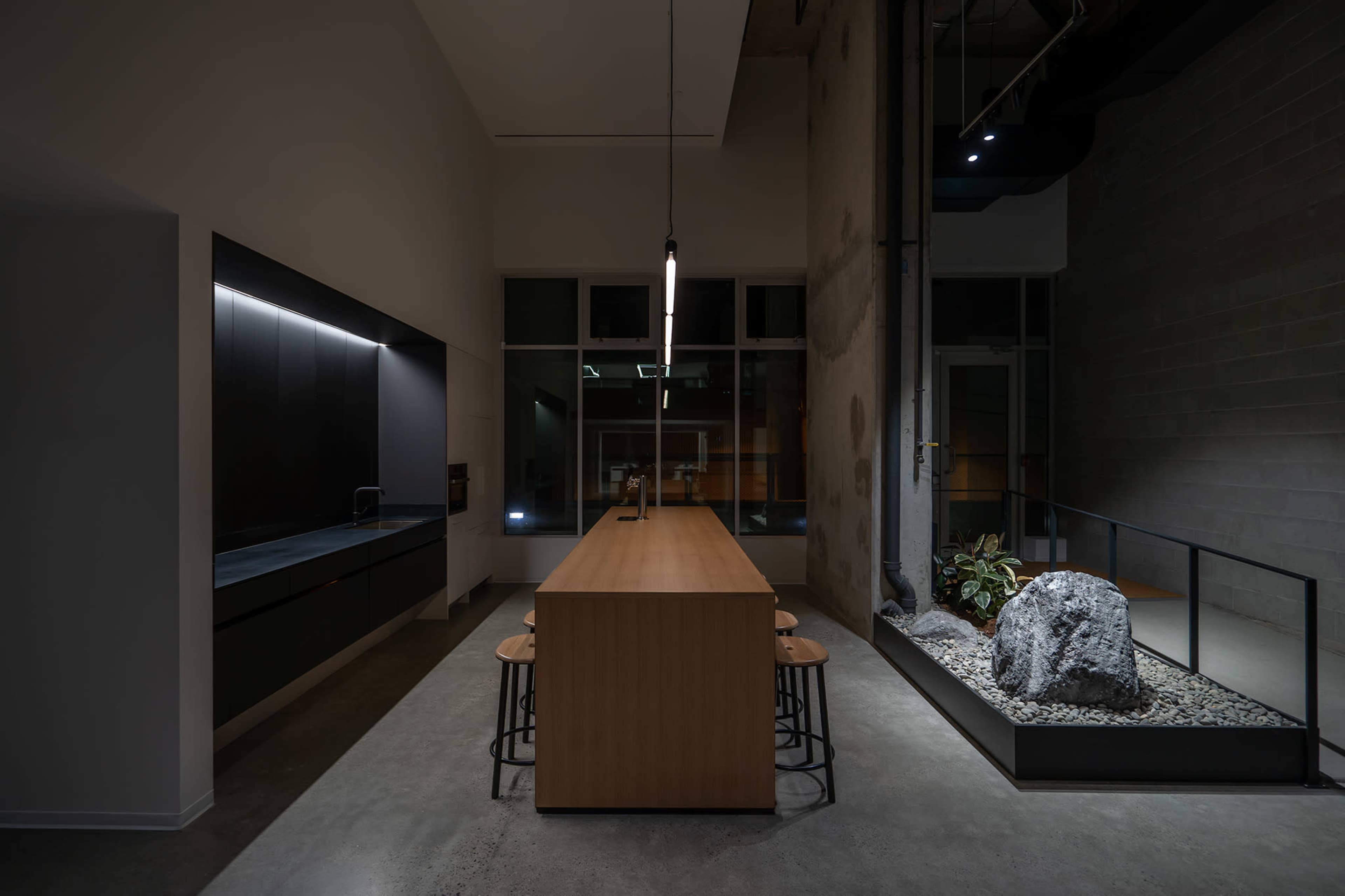 A modern kitchen with a long wooden dining table surrounded by stools, a minimalist design featuring concrete walls, and a rock garden illuminated by soft lighting.