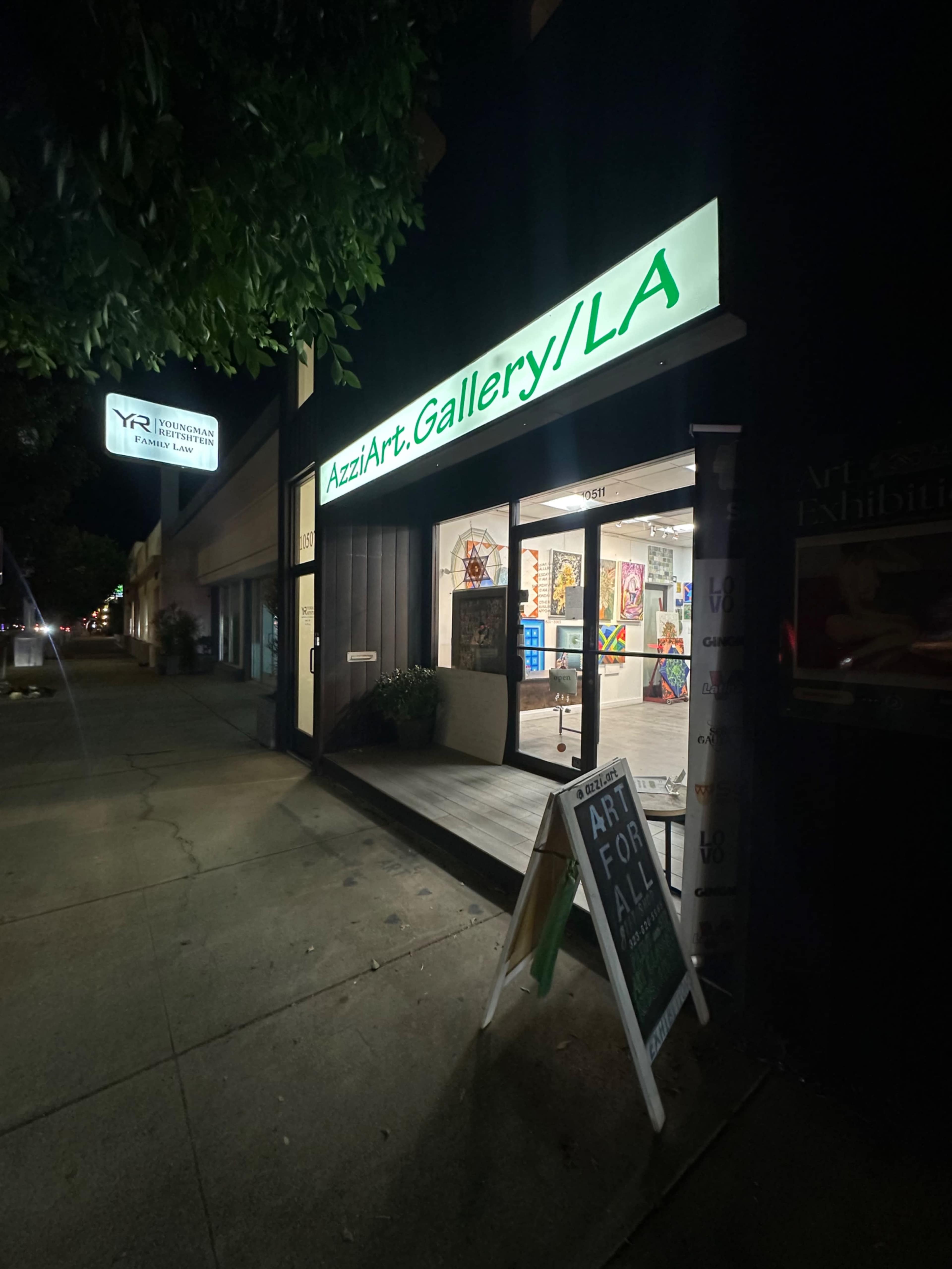 The image shows a brightly lit art gallery entrance at night, featuring a sign that reads "AzziArt.Gallery/LA" and an A-frame sign that says "ART FOR ALL" displayed outside.