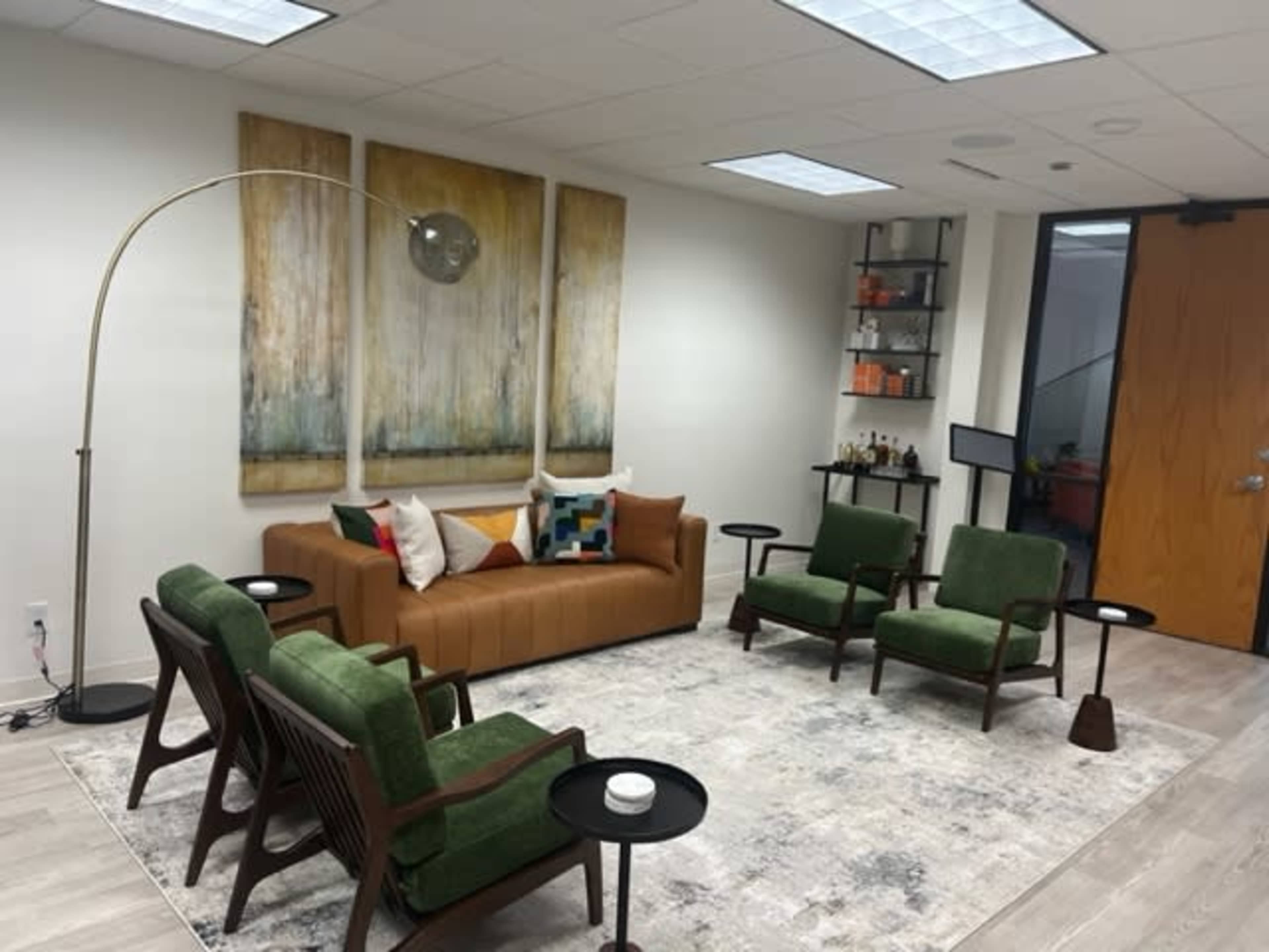 The image shows a modern waiting area featuring a brown sofa, four green chairs, a coffee table, and a large piece of abstract art on the wall.