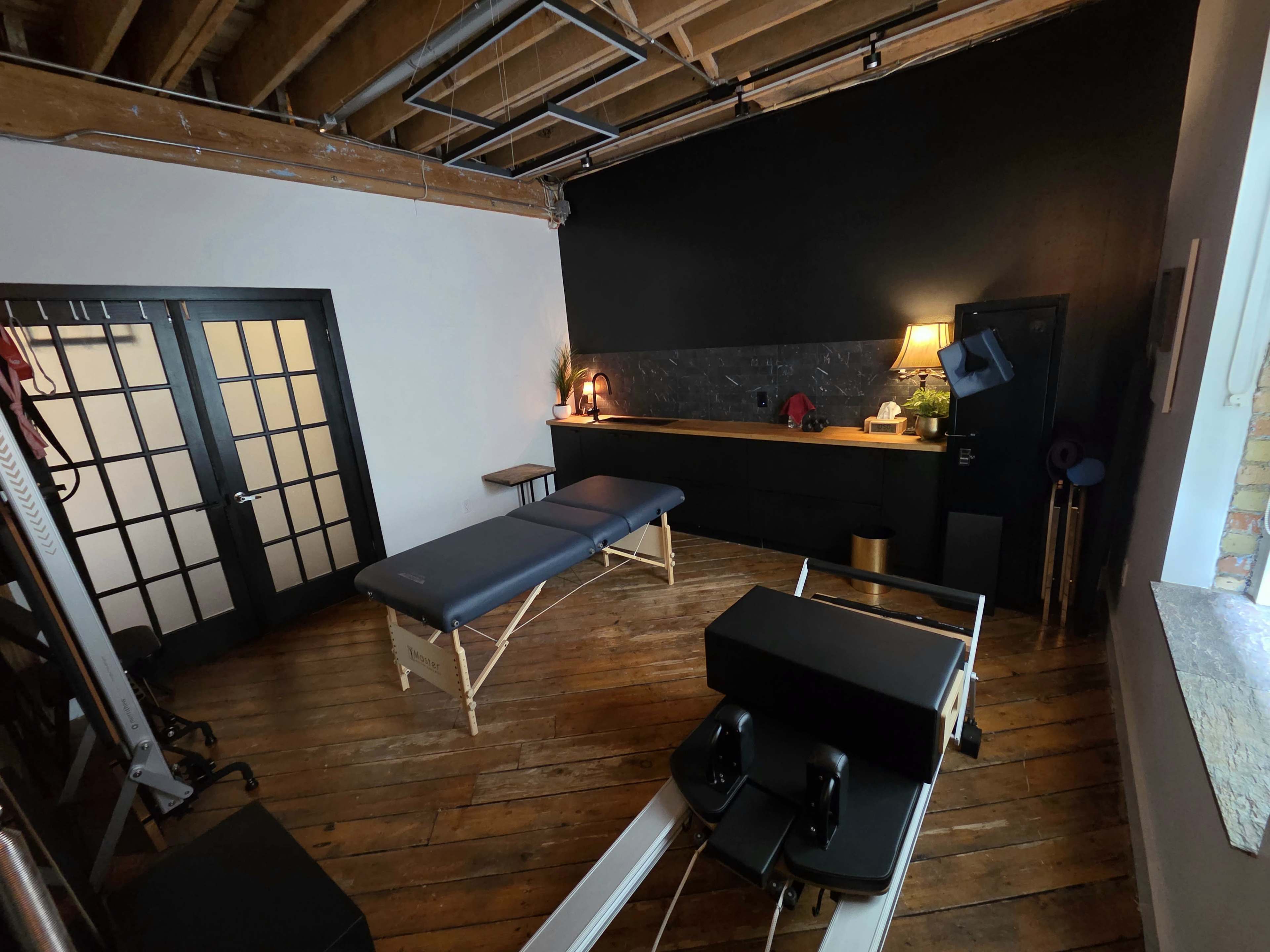 The image shows a fitness studio with a massage table, dark walls, wooden floors, and a reception area featuring a lamp and furniture.