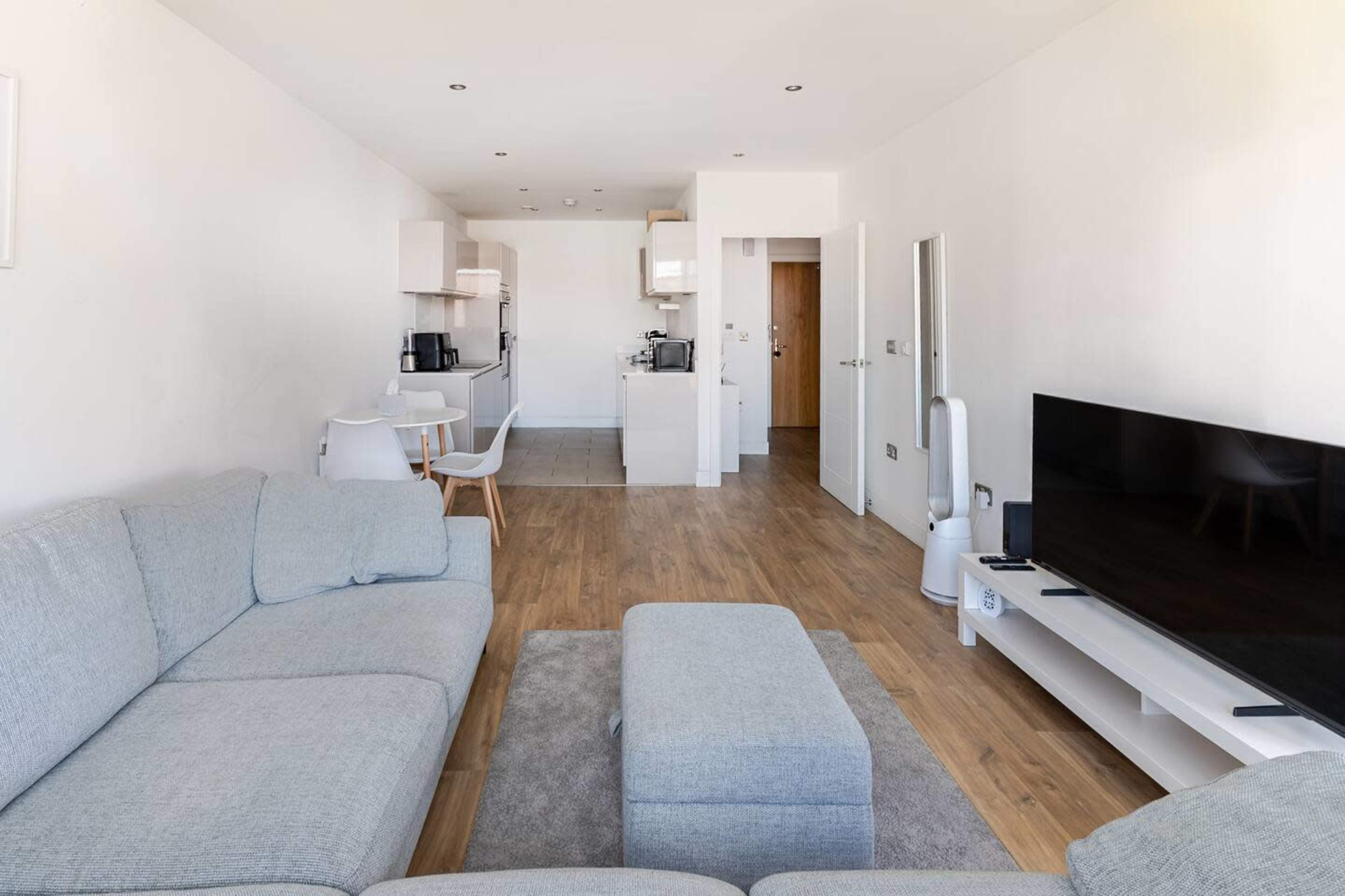 The image shows a modern, open-concept living space featuring a light gray sectional sofa, a coffee table, and a flat-screen TV on a white media console.