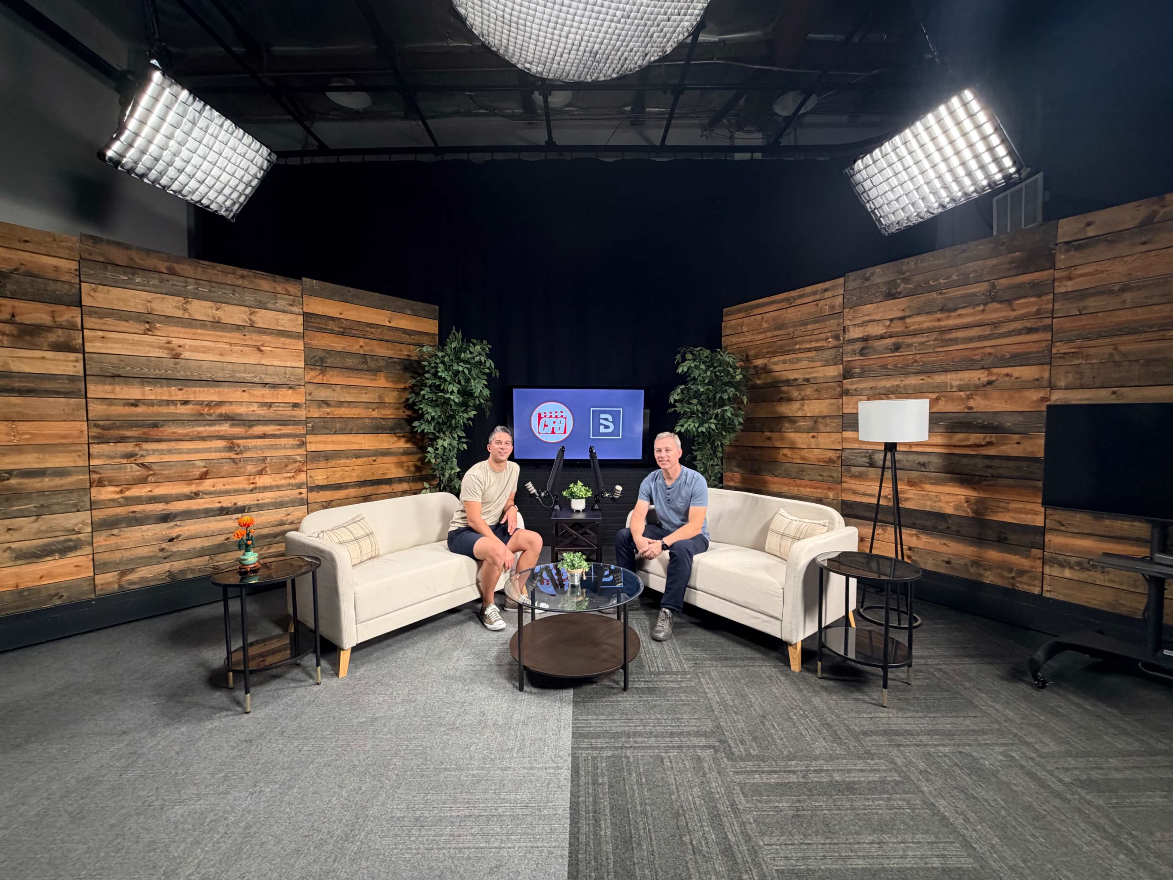 Two men sit on white couches in a studio setting with wooden paneling, surrounded by decorative plants and equipped with studio lighting.
