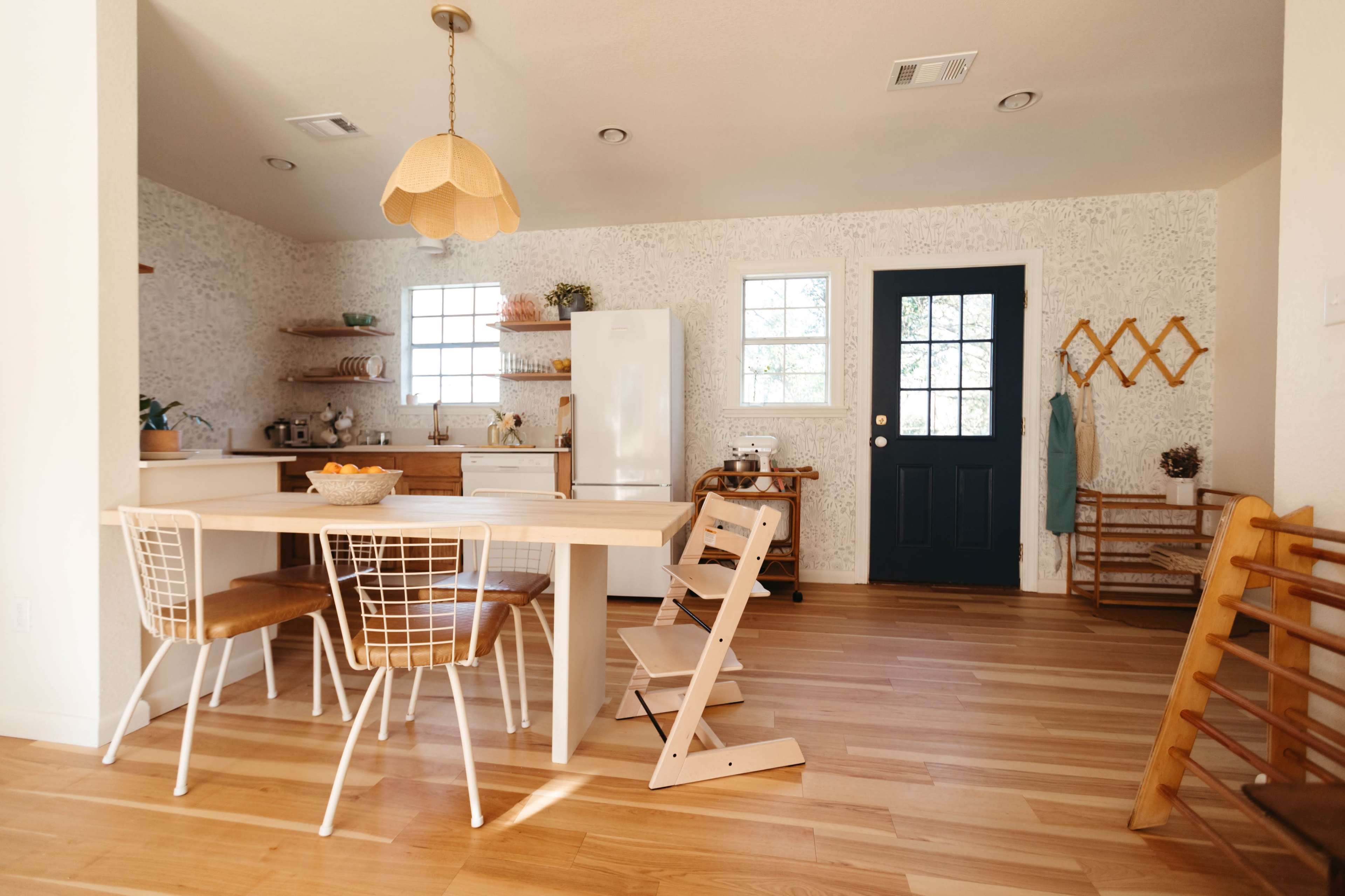 A modern kitchen features a wooden dining table with four chairs, a white refrigerator, and a door leading outside, illuminated by natural light from the windows.