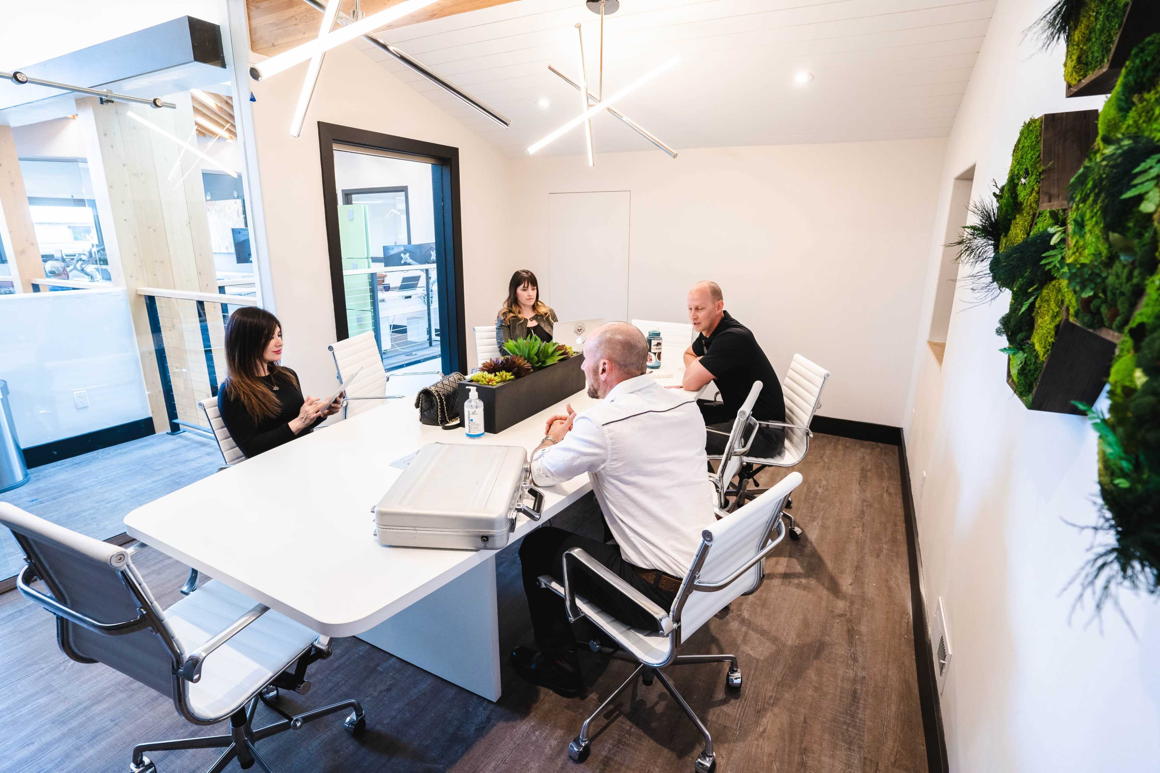 A group of four people is seated around a modern conference table in a well-lit meeting room with green wall art.