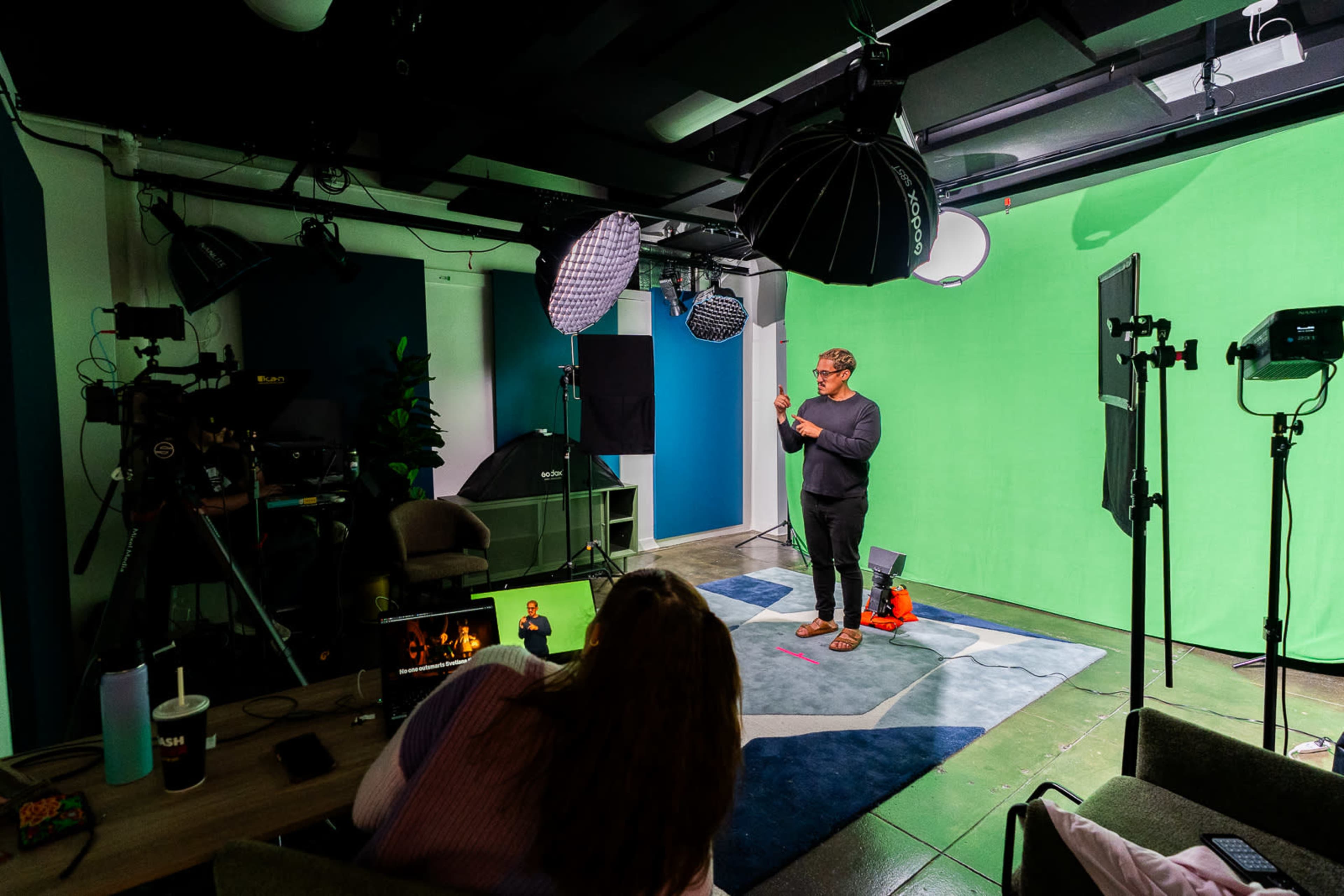 A person stands in front of a green screen studio while cameras and lighting equipment are set up around the room.
