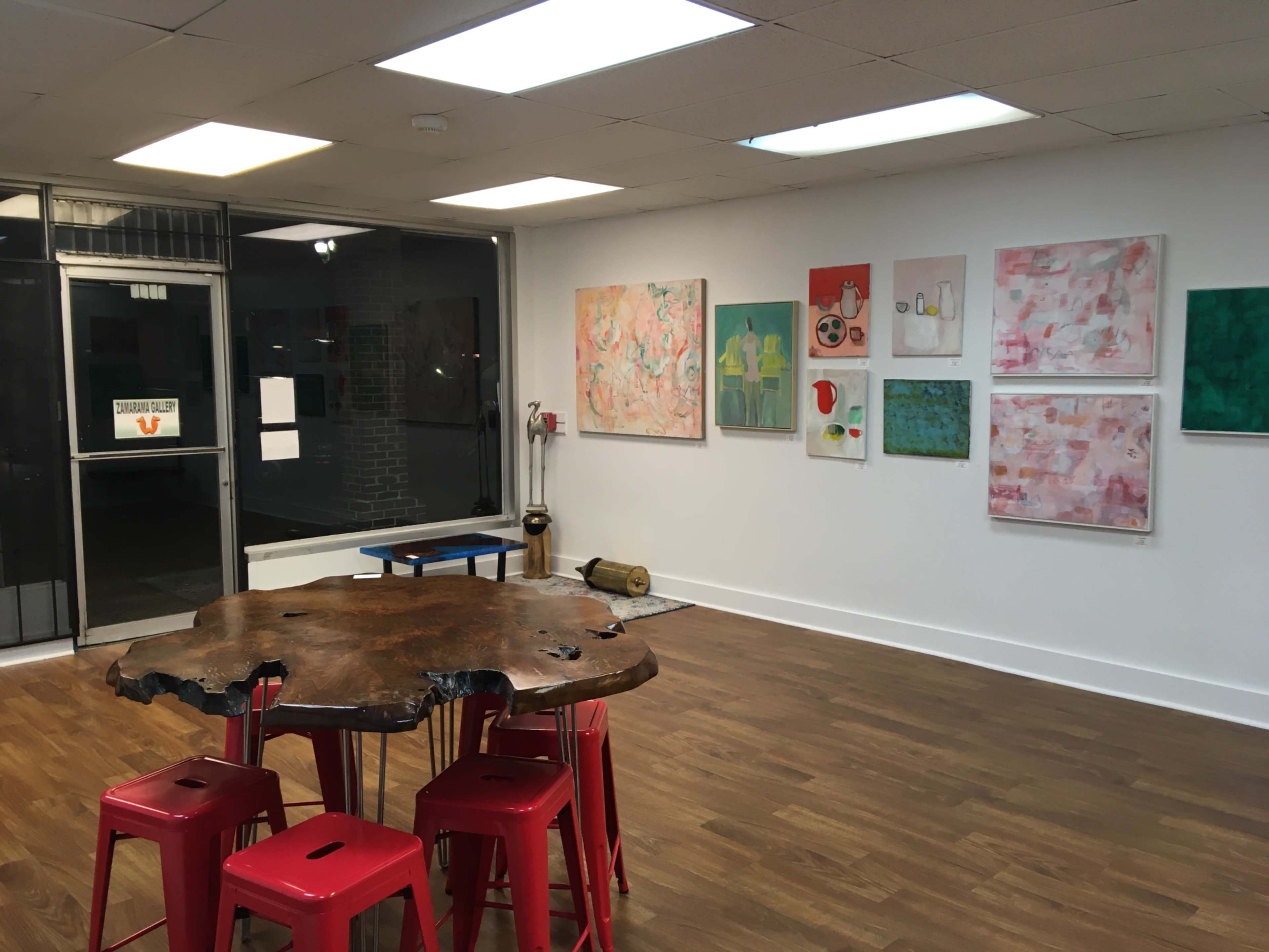 The image shows a gallery space with a large wooden table surrounded by red stools, and colorful abstract paintings arranged on the white walls.