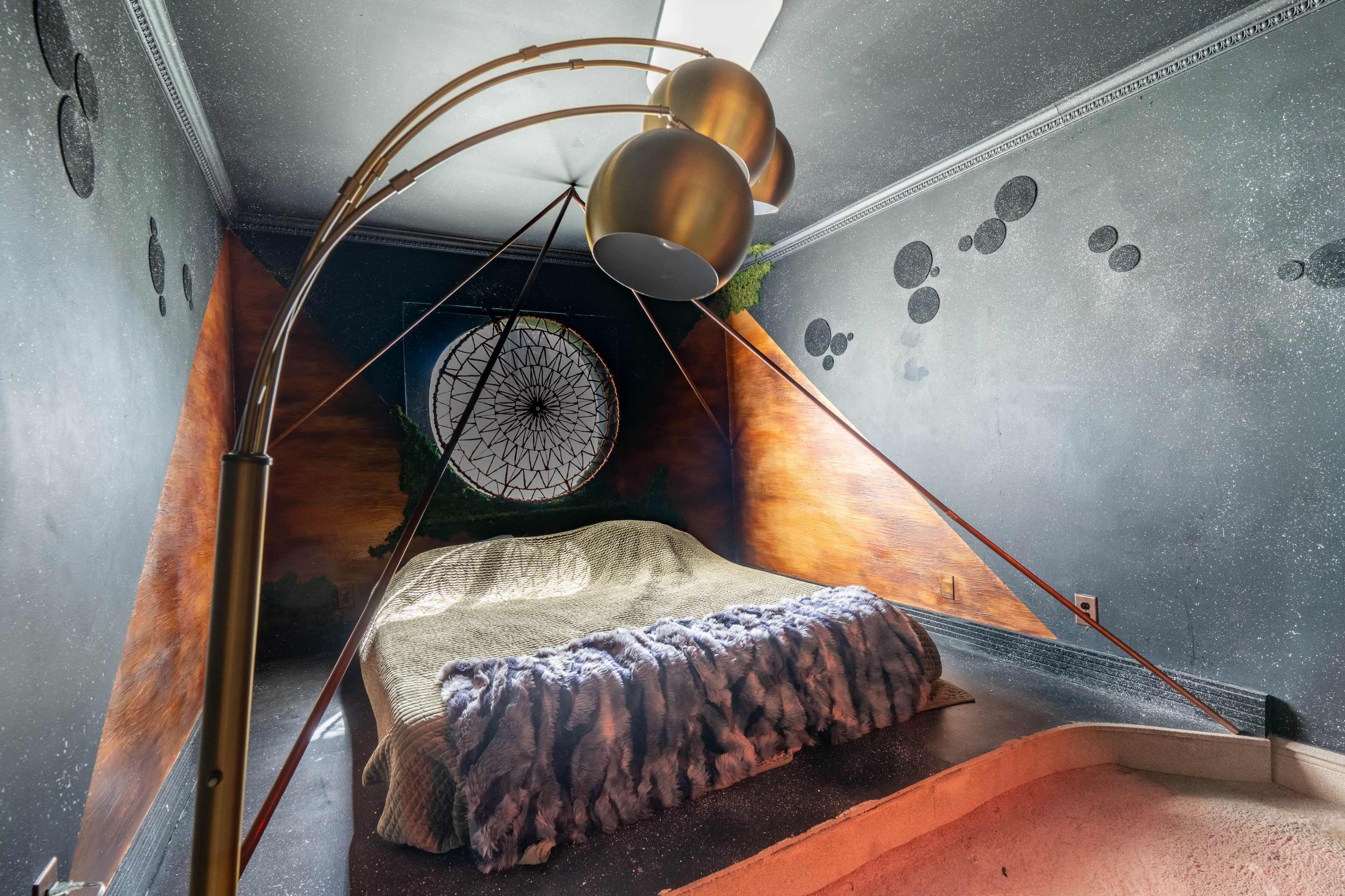 The image shows a creatively designed bedroom featuring a bed with a textured blanket, a unique floor lamp, and artistic wall murals.
