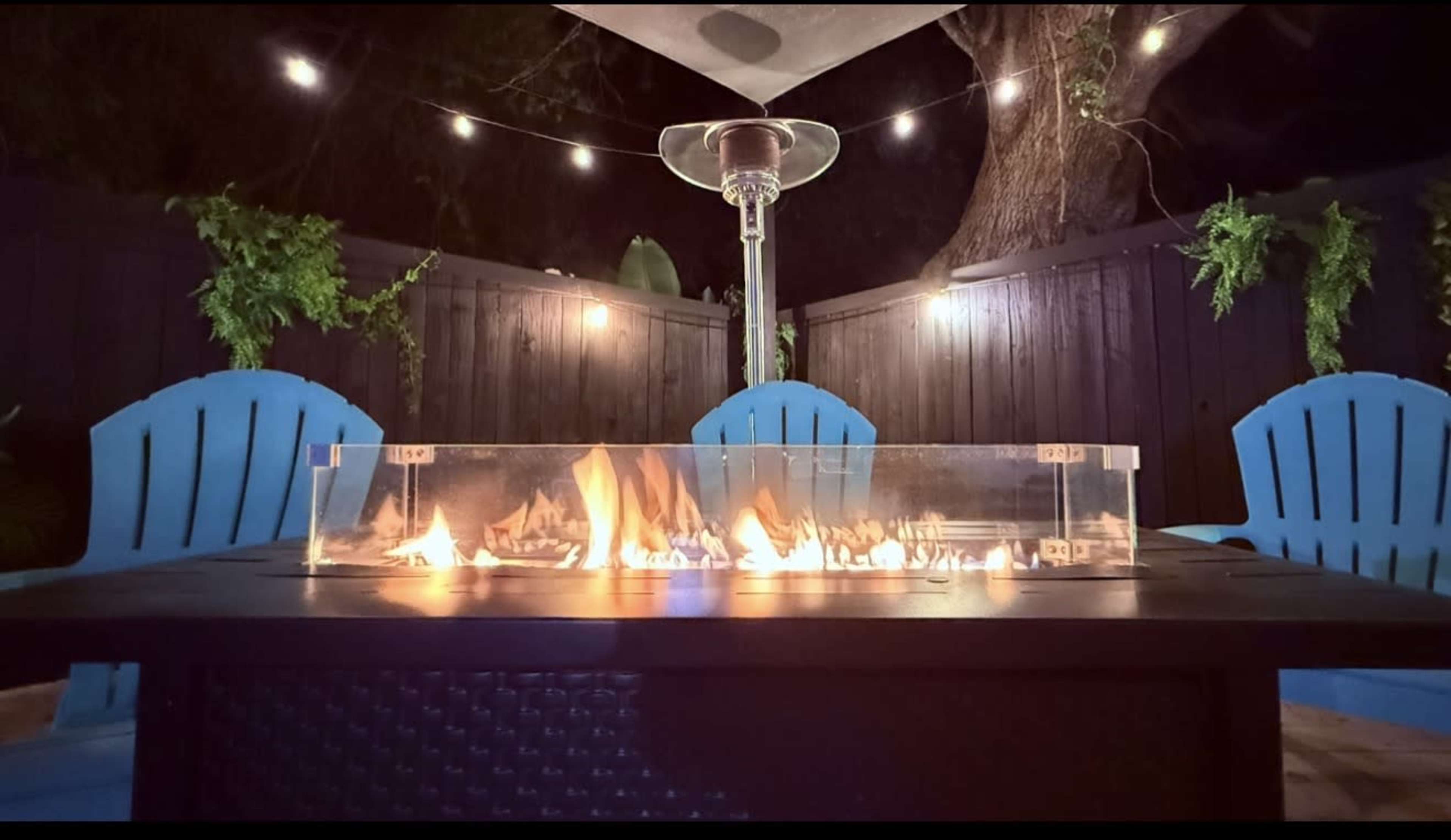 A modern outdoor seating area features a table with a glass fire pit surrounded by blue chairs and illuminated by string lights above.