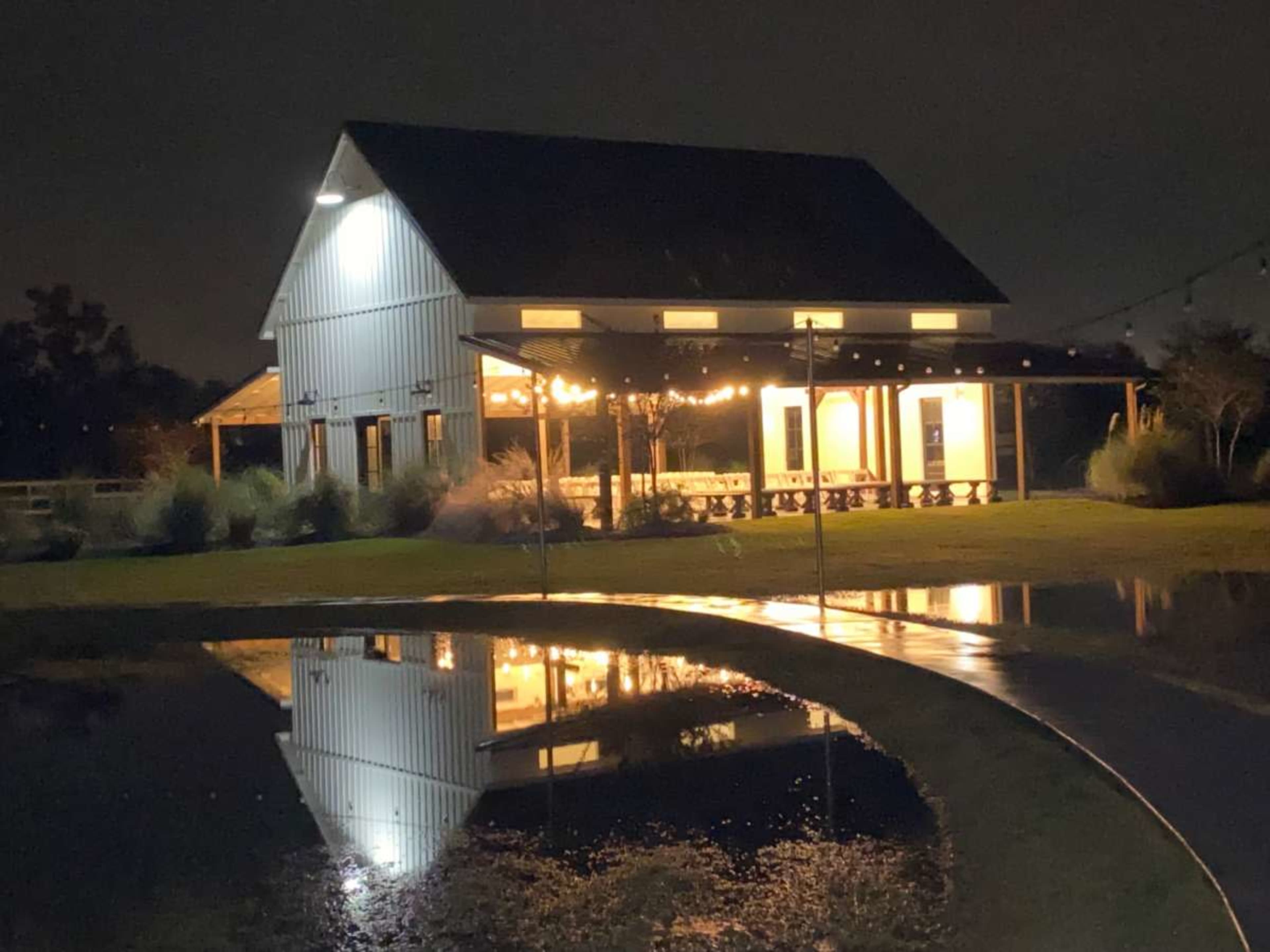 A modern barn with lighted windows stands at night, reflecting in a nearby puddle on the ground.