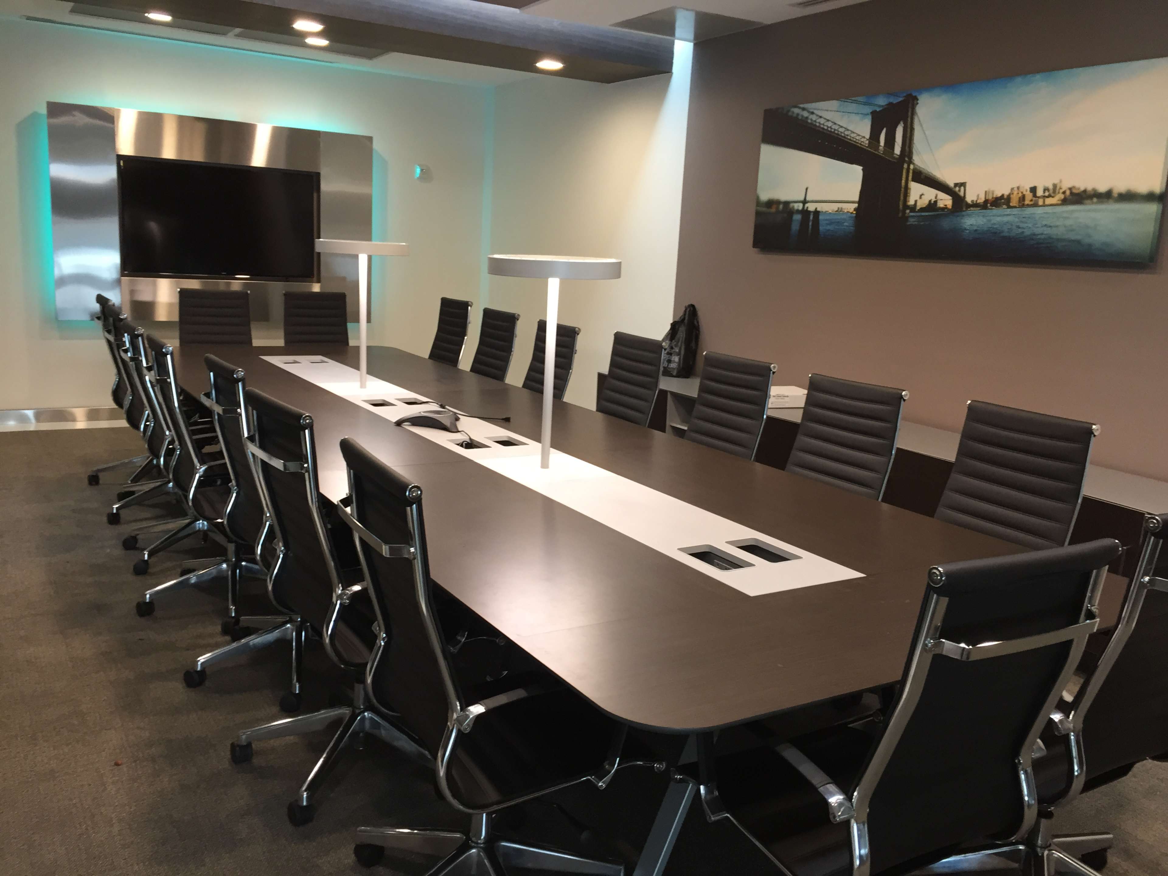 A modern conference room featuring a long table surrounded by black chairs, with a large screen on one wall and a cityscape artwork on another.