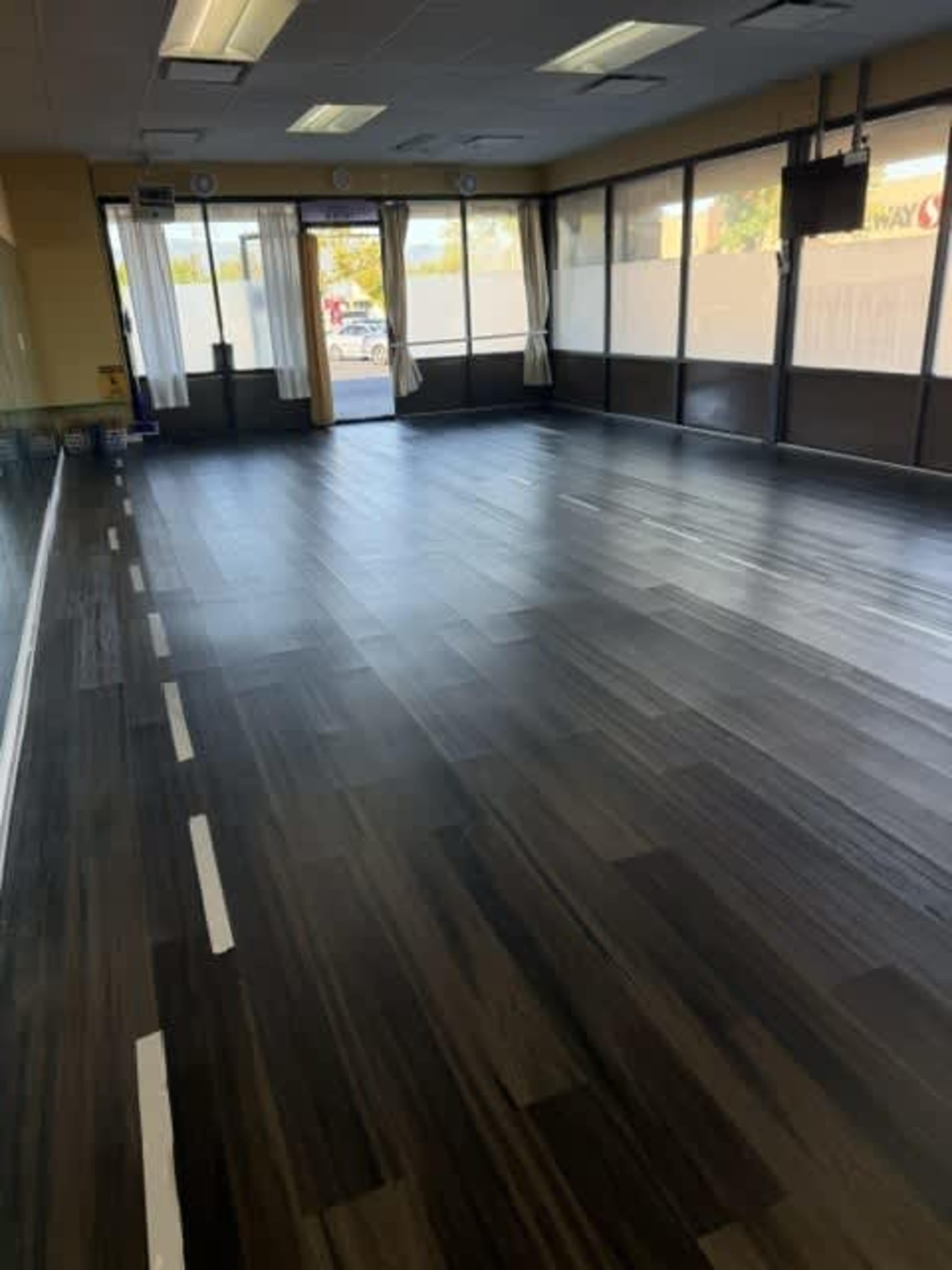 The image shows a spacious indoor area with dark wood-like flooring, large windows, and a few curtains.