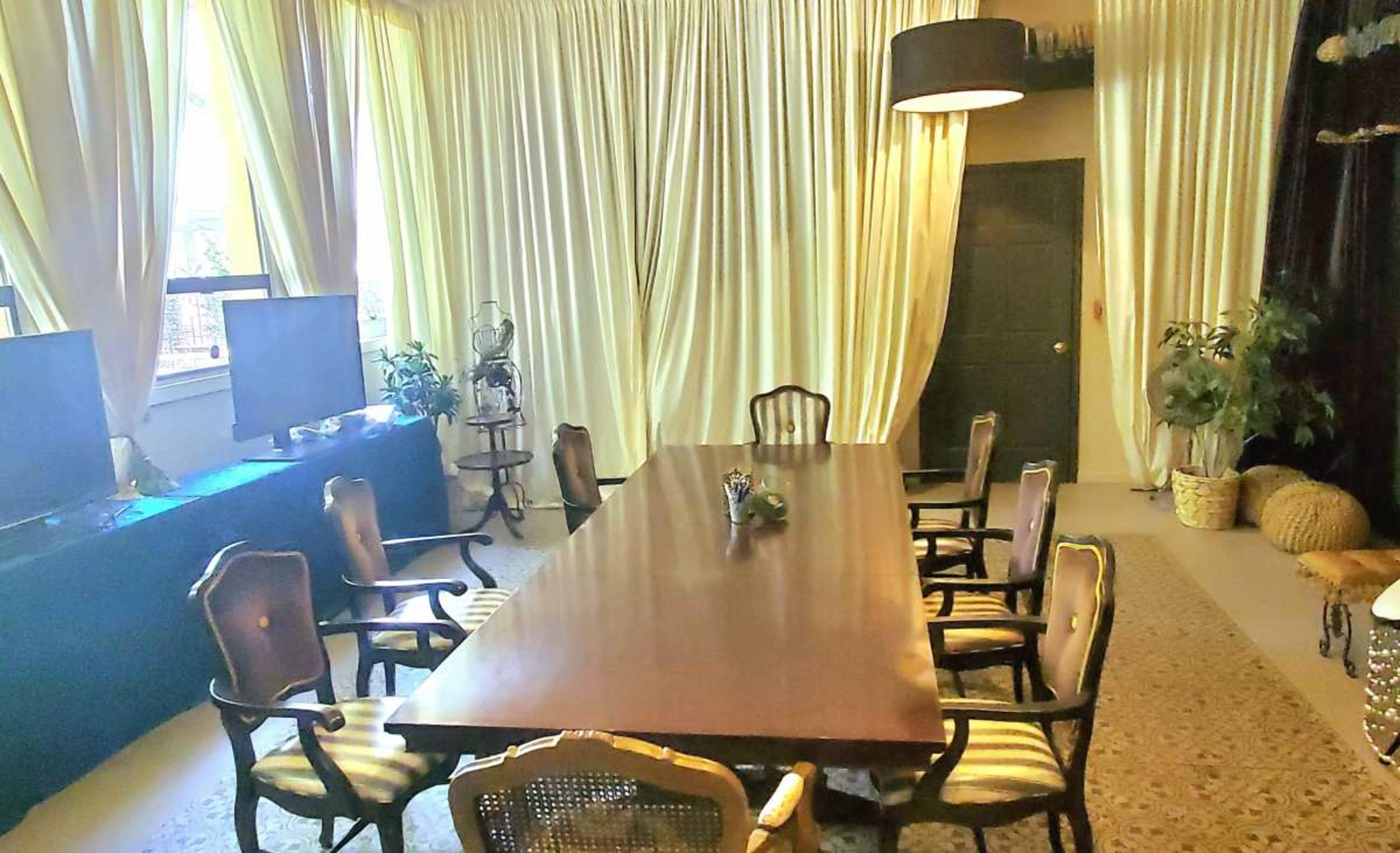 A long wooden table surrounded by upholstered chairs is set in a room with yellow curtains and two large television screens on a black cabinet.
