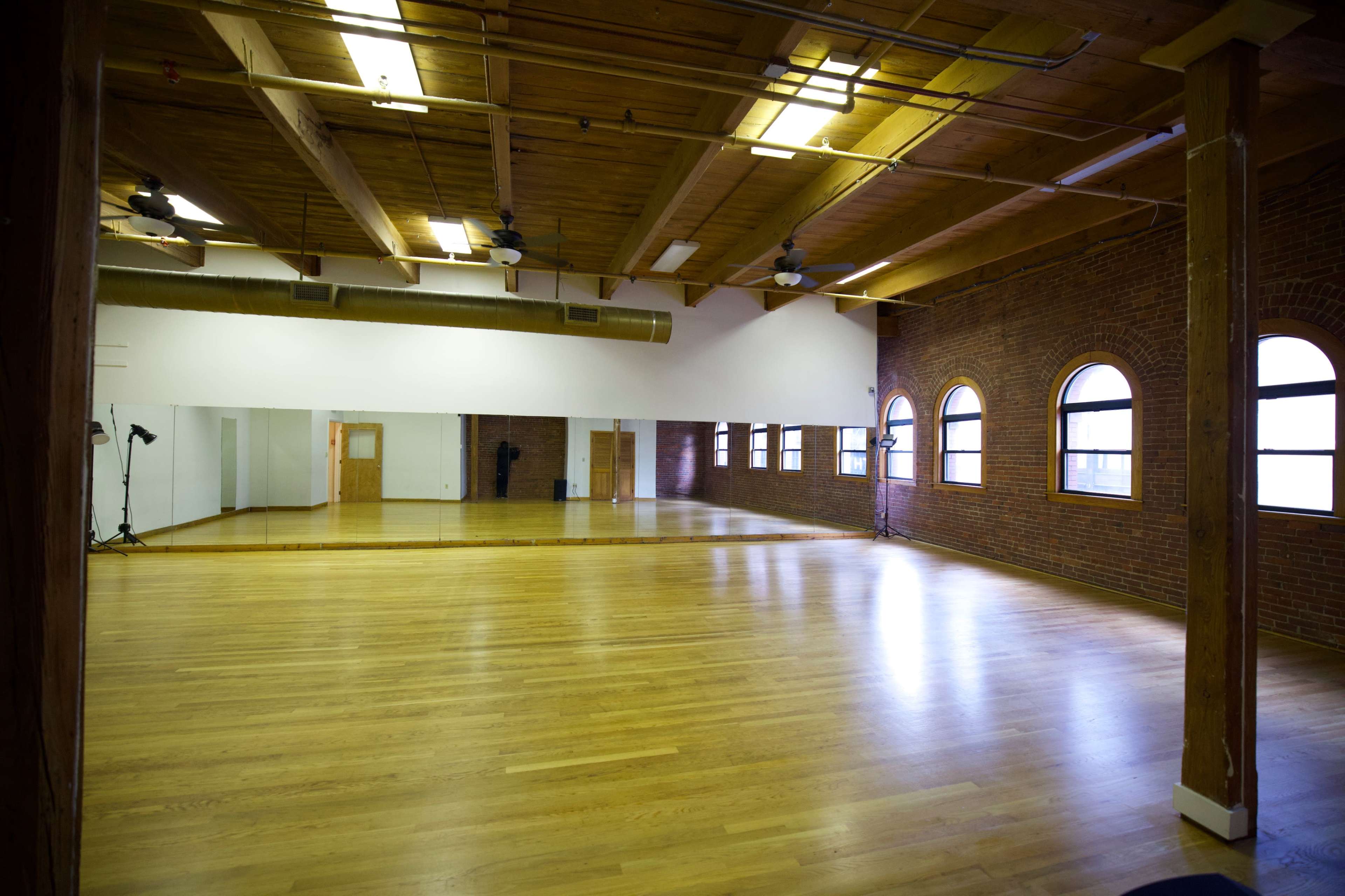 The image shows an empty dance studio with wooden floors, exposed beams, and large arched windows.