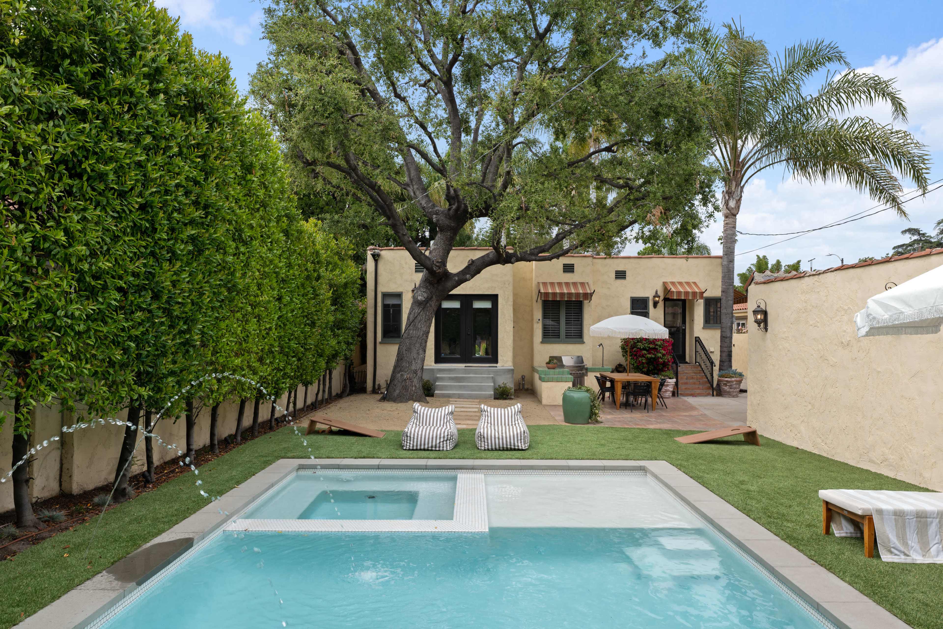 The image shows a backyard featuring a small pool, two lounge chairs, and a large tree alongside a house with a patio and outdoor dining area.