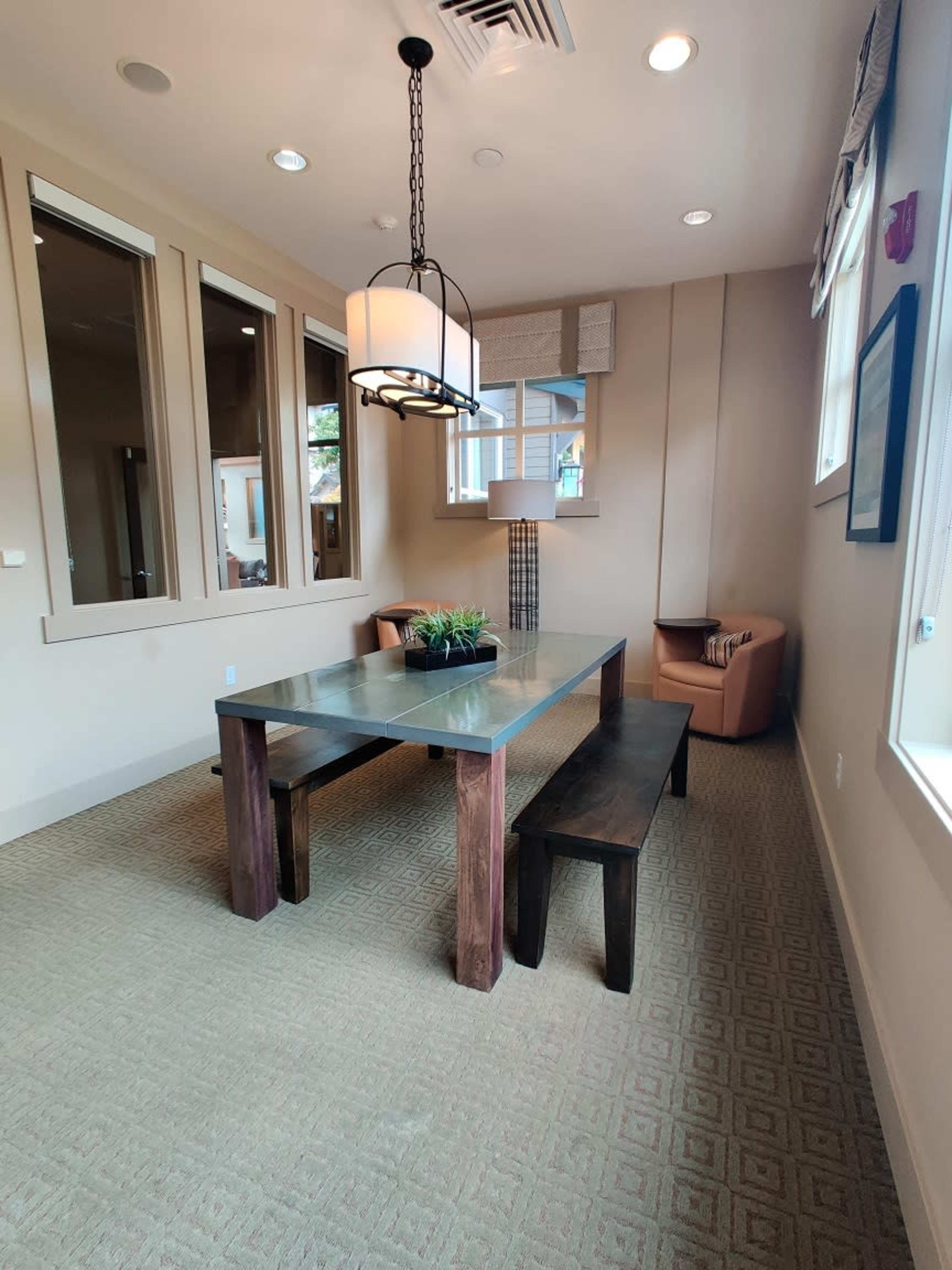 The image shows a small meeting room furnished with a rectangular table, two benches, a pendant light, and a corner chair, all set against a neutral-colored wall with windows.