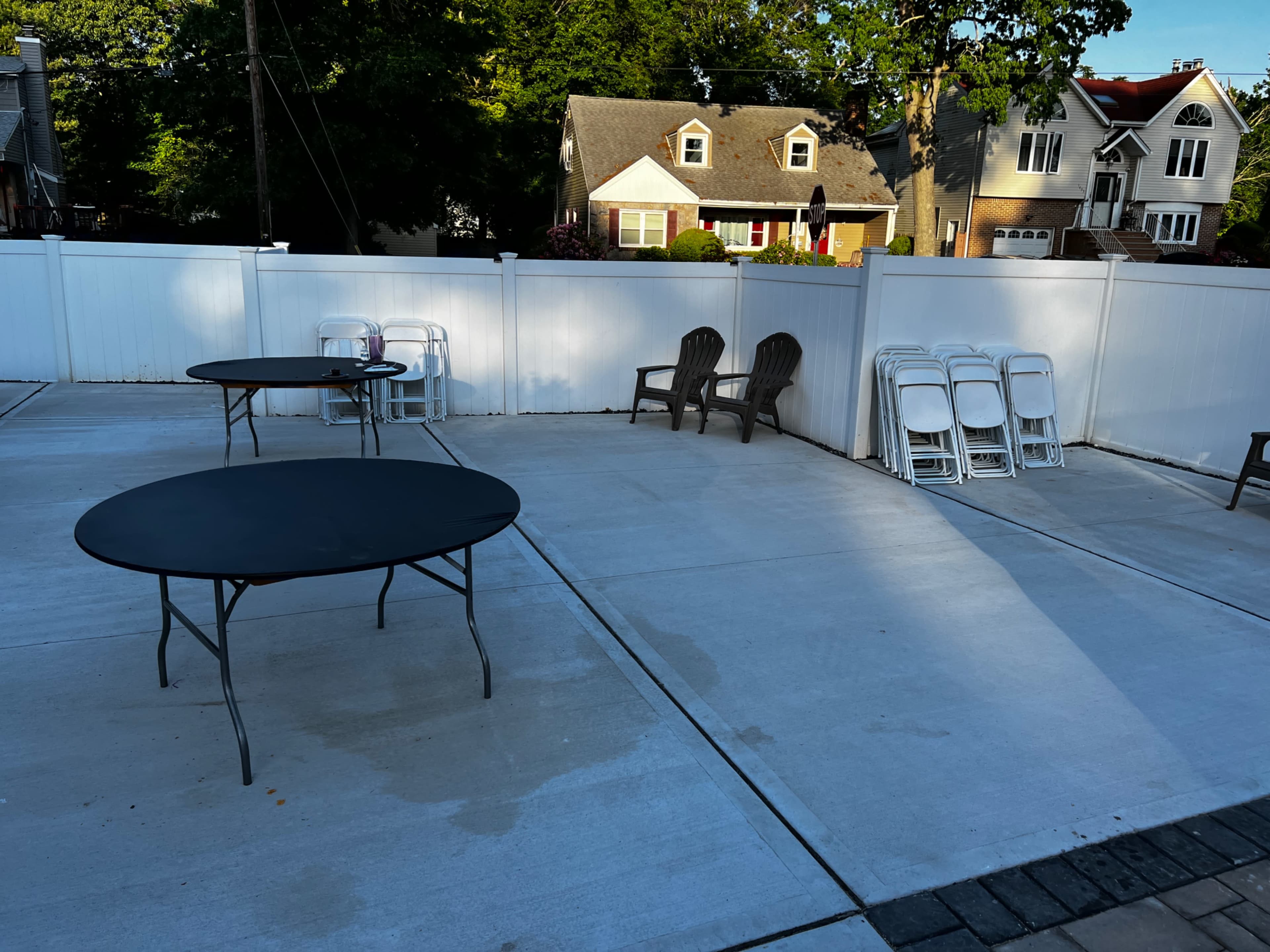 The image shows a fenced patio area with two round tables, a few outdoor chairs, and a stack of folding chairs against the fence.