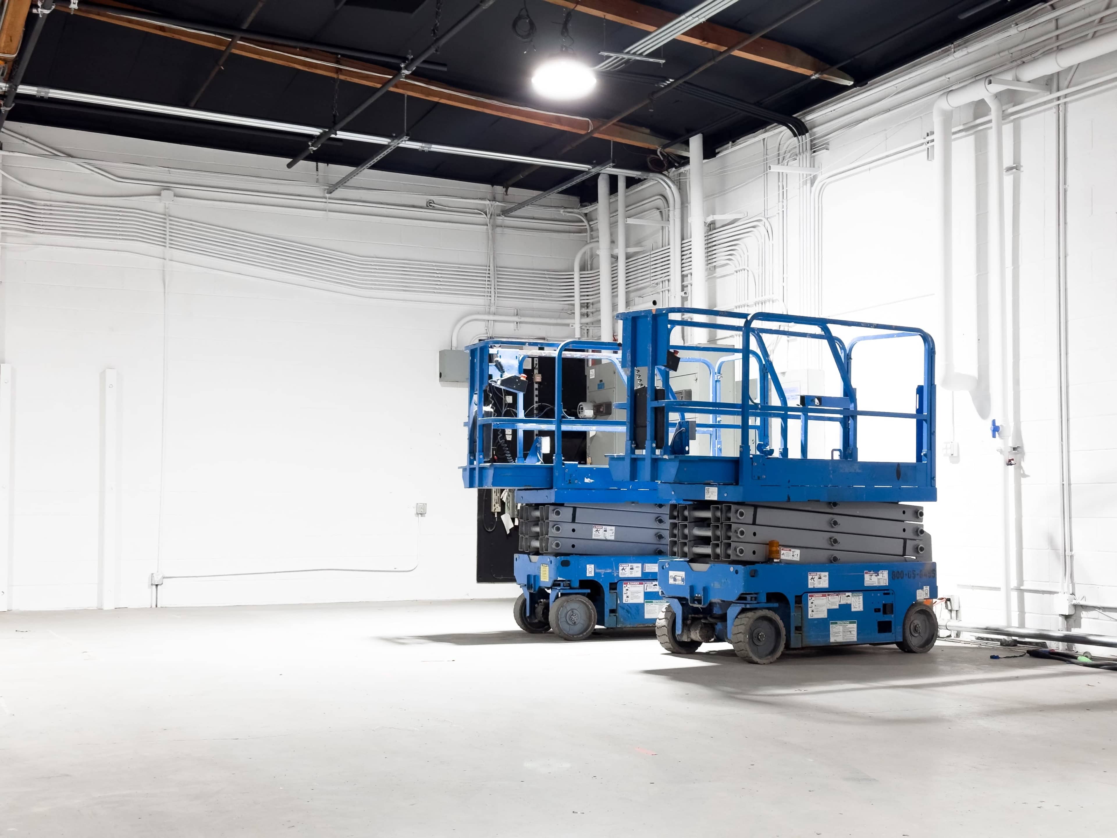 Two blue scissor lifts are parked on a concrete floor in a spacious industrial room with white walls and exposed piping.