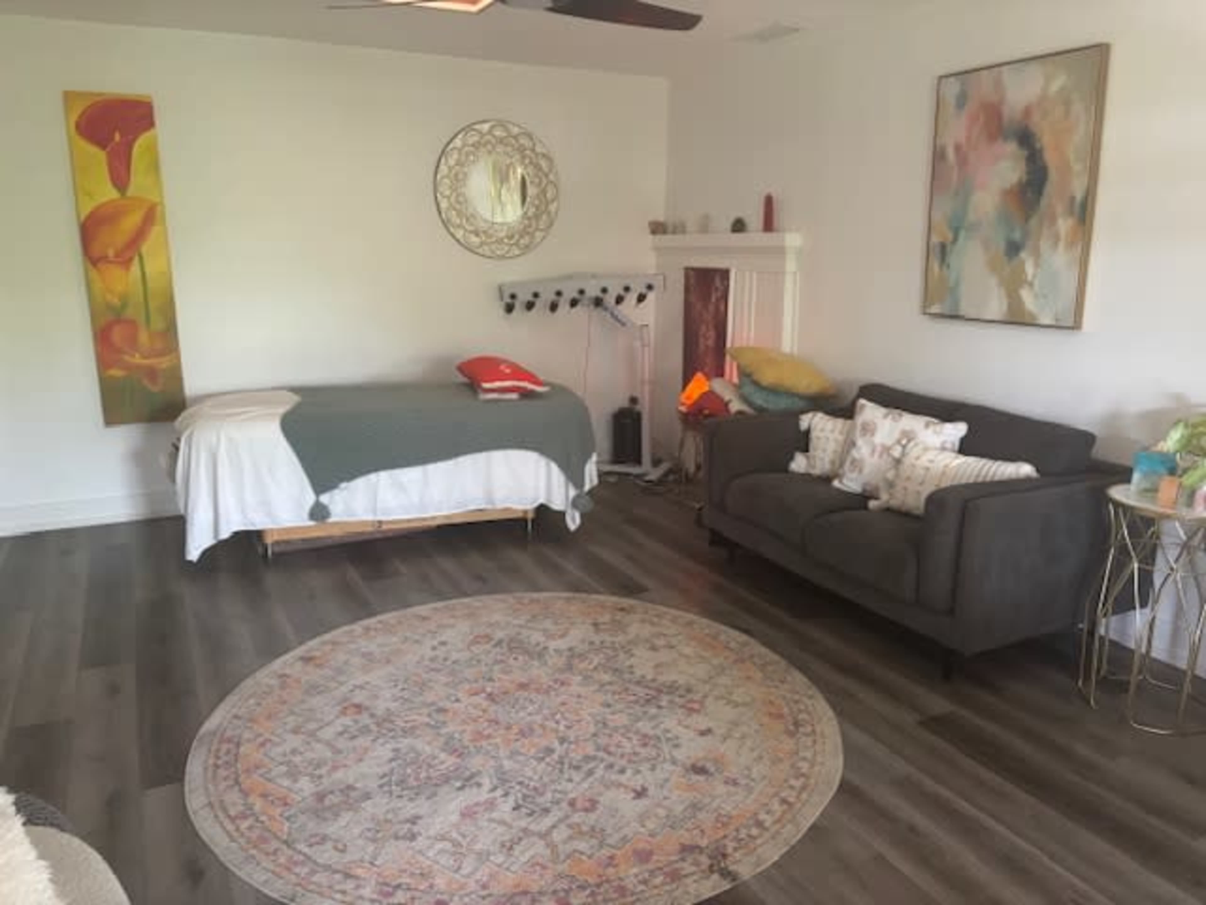 The image shows a cozy room with a massage table, a gray sofa, a round patterned rug, and decorative artwork on the walls.