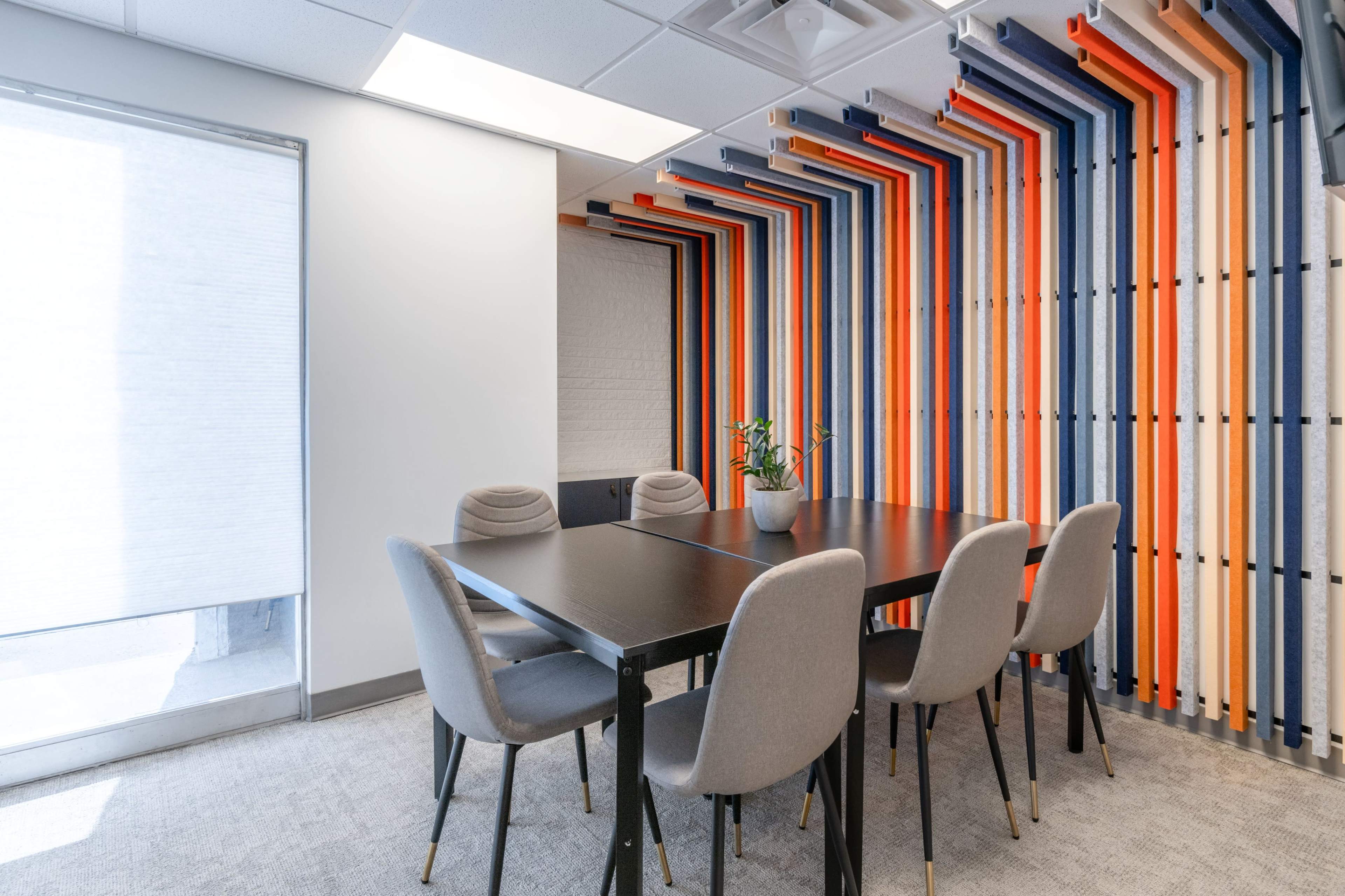 A conference room features a black table surrounded by gray chairs, with a vibrant striped wall in shades of blue, orange, and white.
