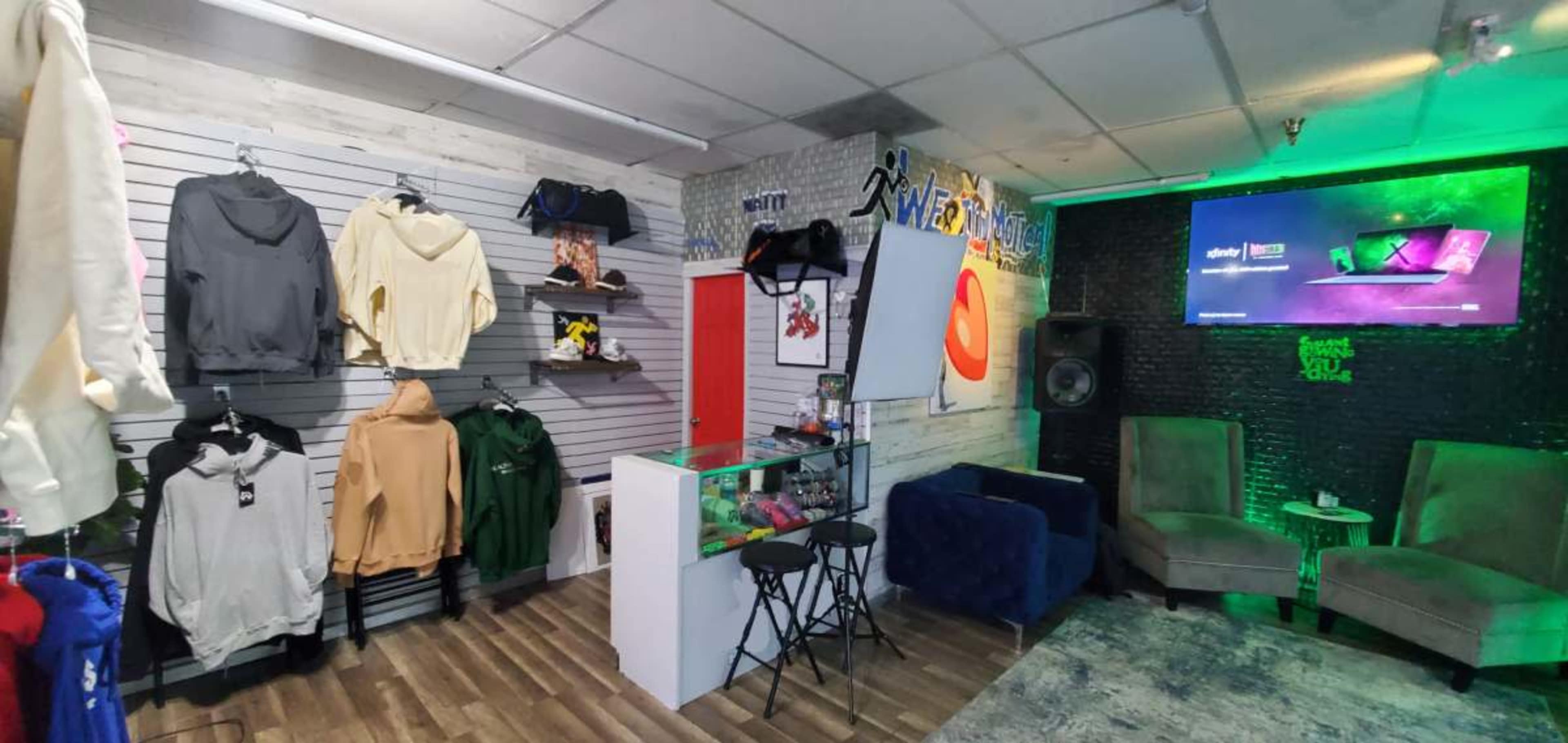 The image shows a retail space featuring clothing displays on the left, a small seating area with two chairs, and a television mounted on the wall.