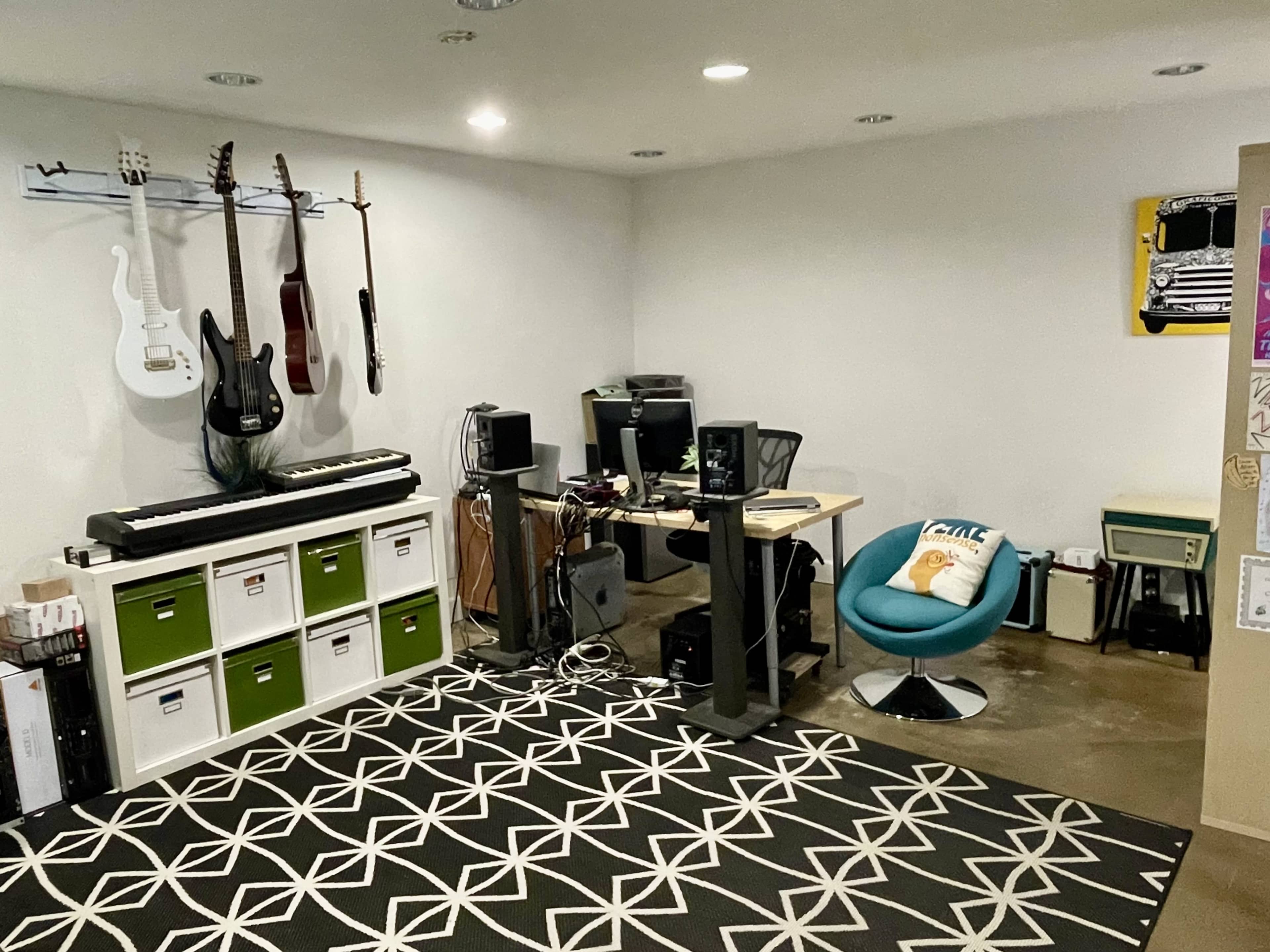 A compact music studio features guitars on the wall, a keyboard, a desk with audio equipment, and a blue chair on a patterned rug.