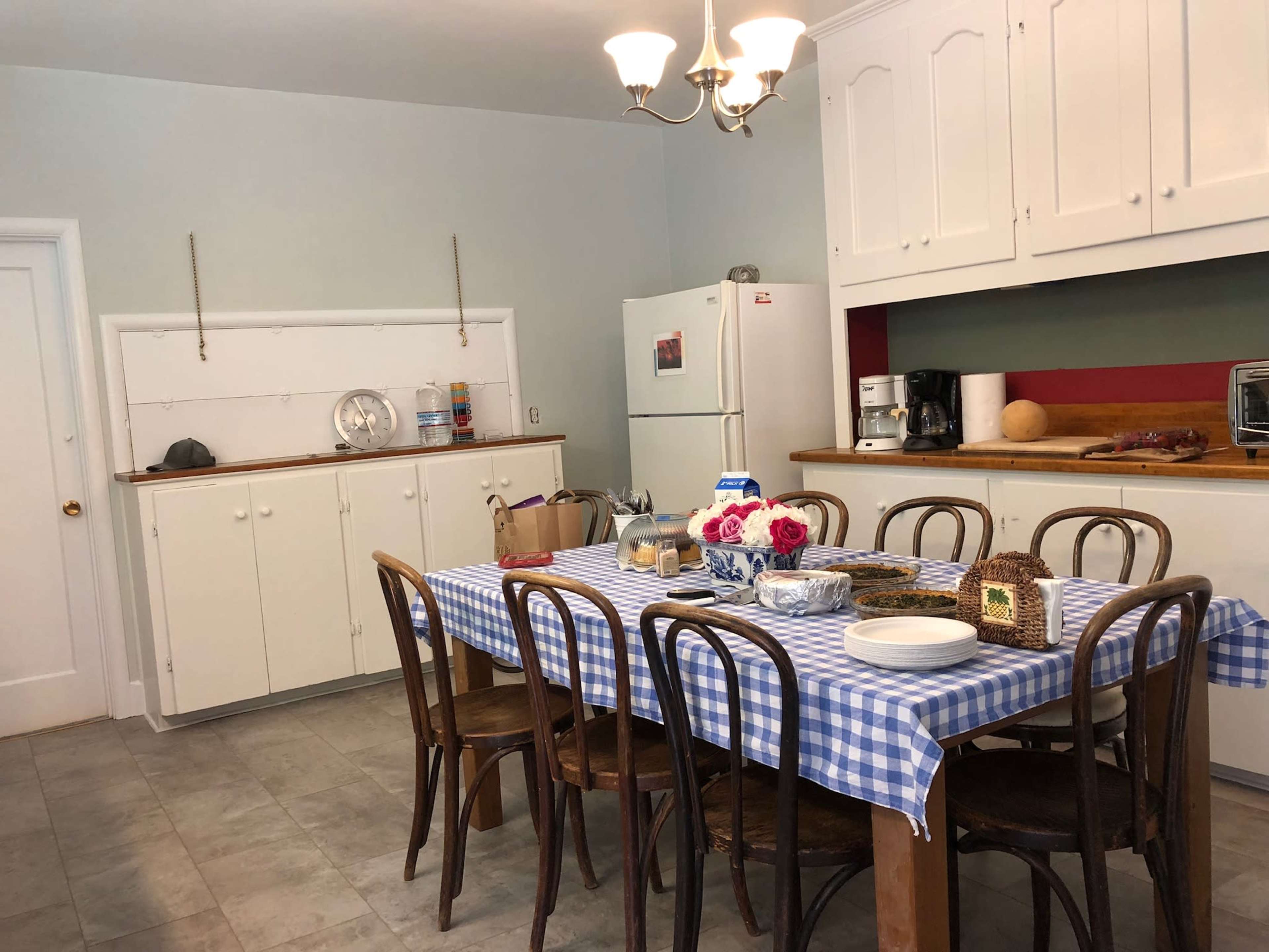 A kitchen features a checkered tablecloth-covered dining table surrounded by wooden chairs, with white cabinets and various appliances in the background.