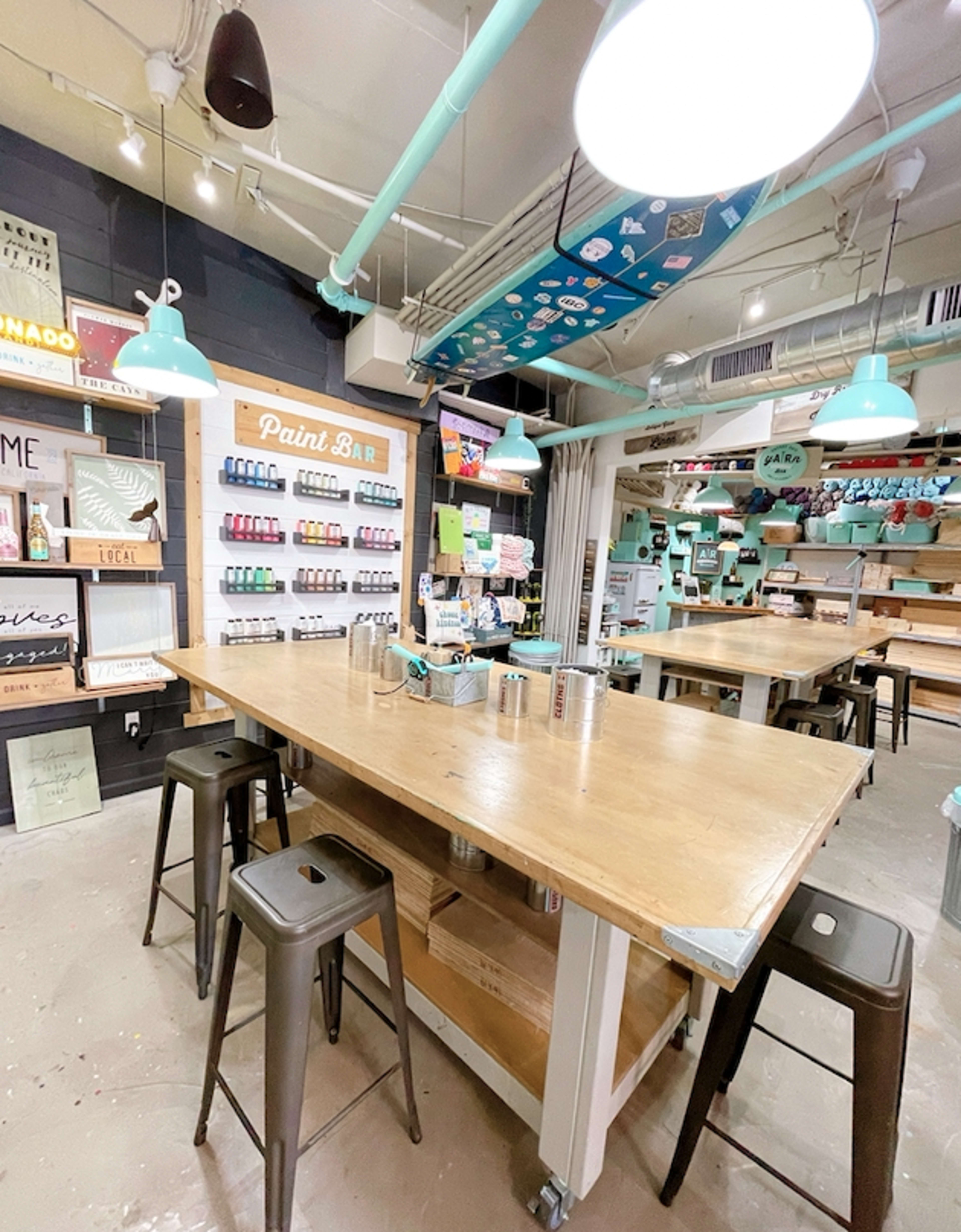A brightly lit art studio with several wooden tables, teal pendant lights, and shelves displaying an assortment of paint supplies.