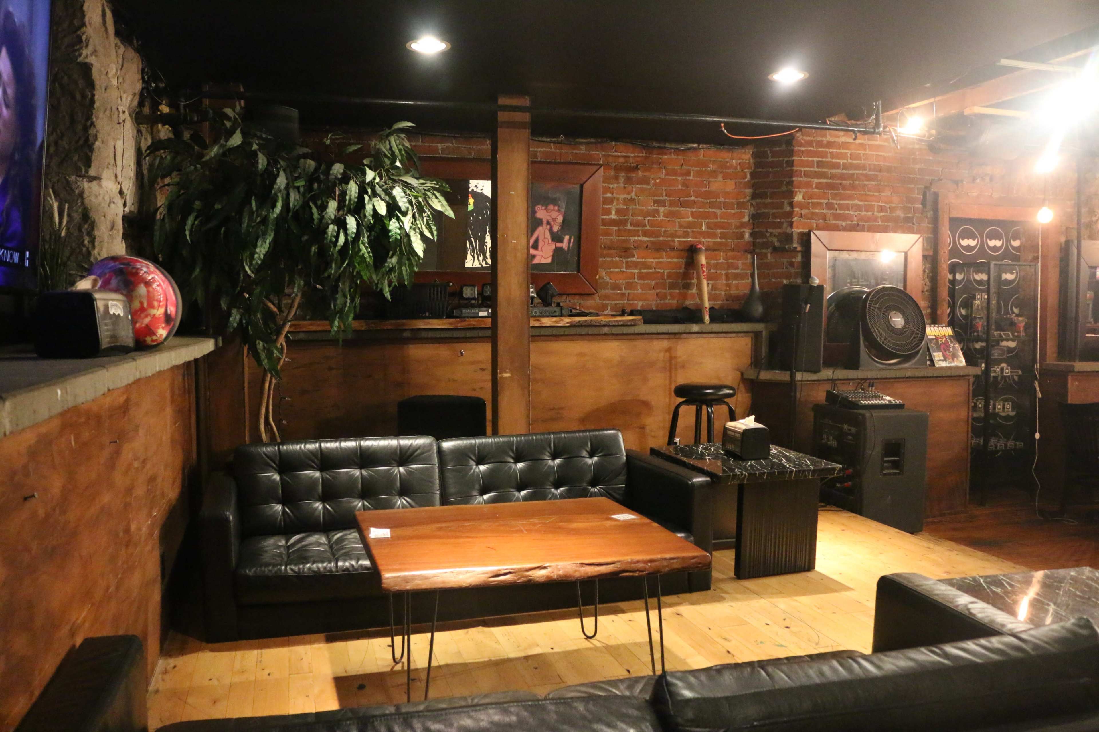 A dimly lit lounge area featuring leather sofas, a wooden coffee table, and a bar with exposed brick walls.