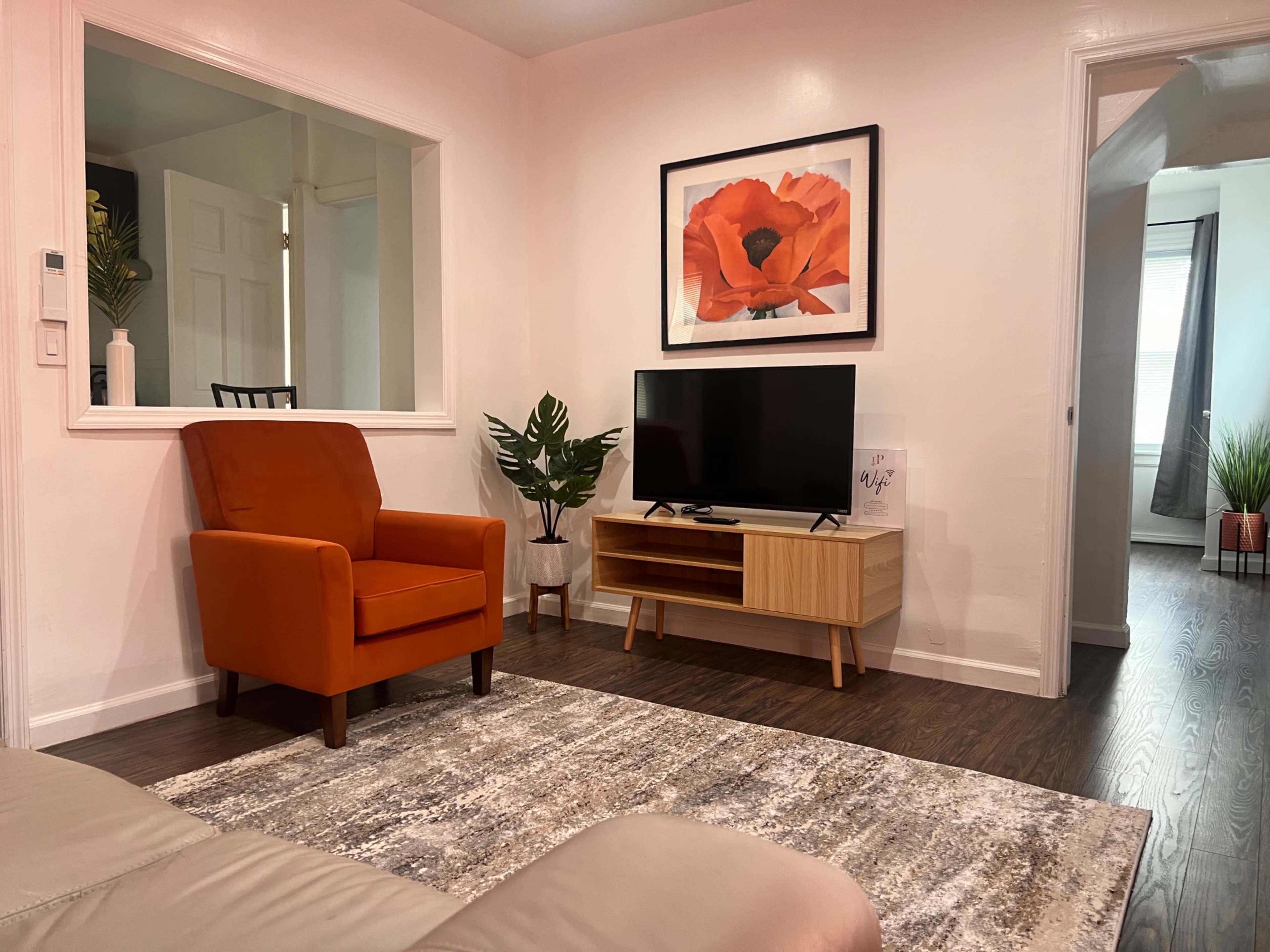 A cozy living room features an orange armchair, a wooden TV stand with a television, and a vibrant flower painting on the wall, with a plant in the corner.